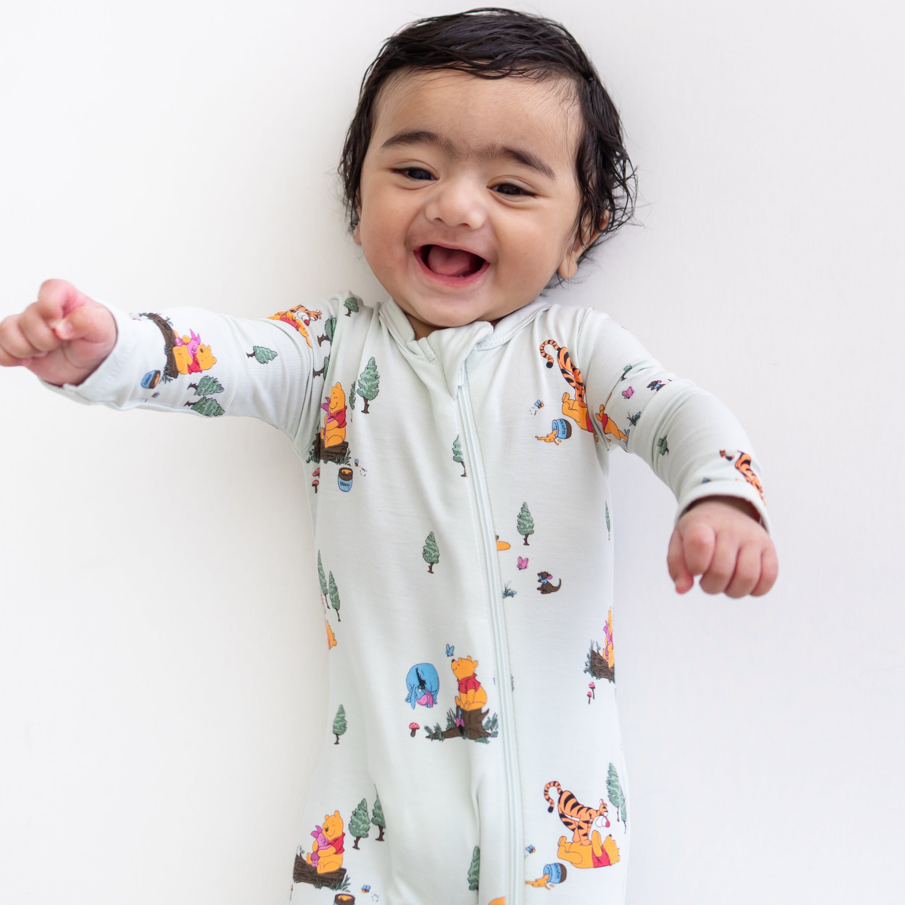 Close up of smiling model wearing the Zippered Romper in Winnie the Pooh Hundred Acre Wood laying on a white surface