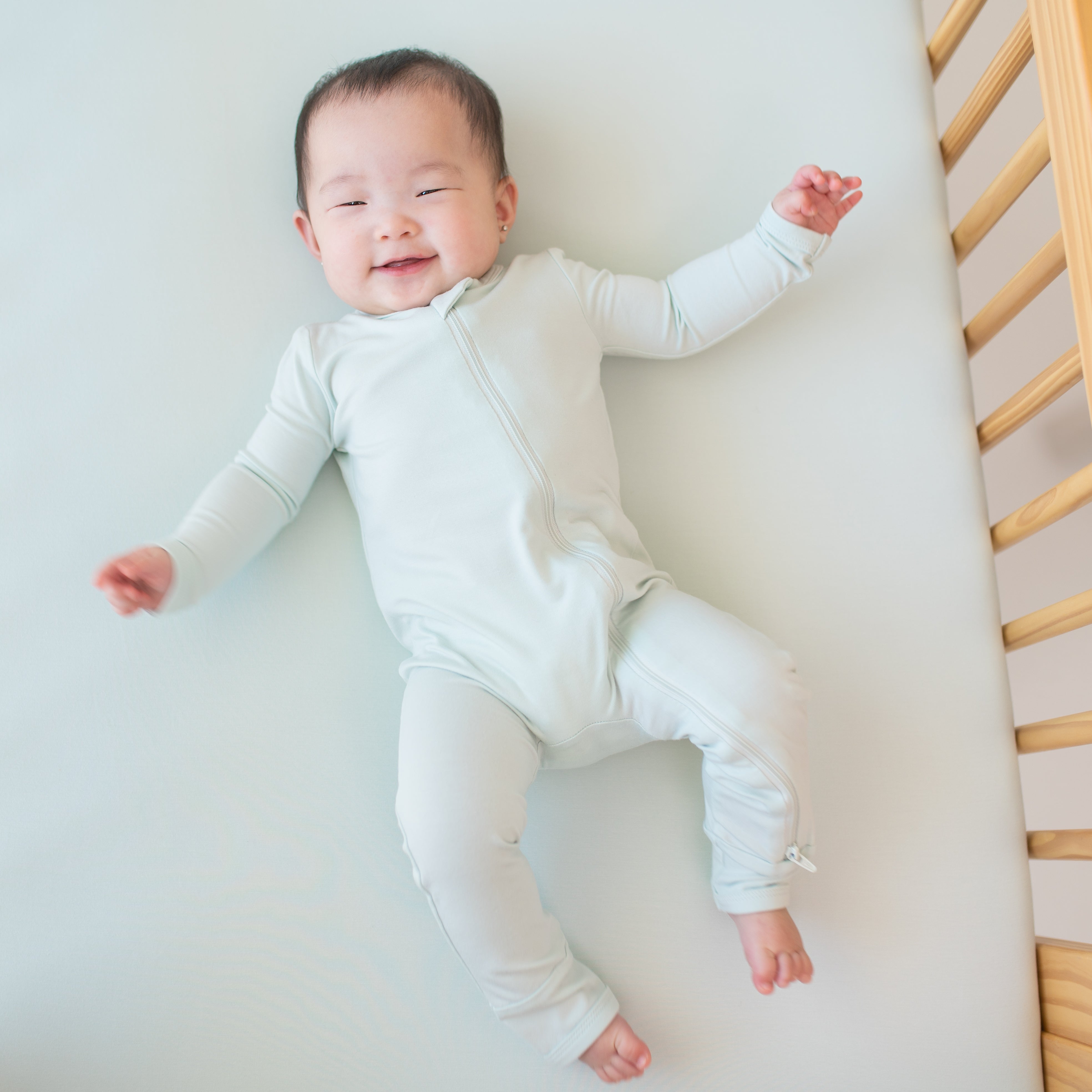 Smiling infant laying in crib wearing the Zippered Romper in Dew