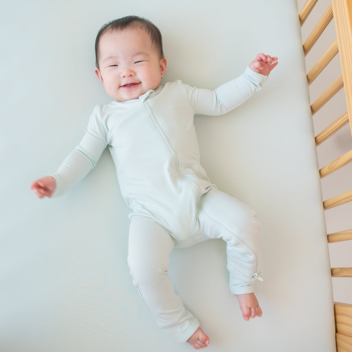 Smiling infant laying in crib wearing the Zippered Romper in Dew