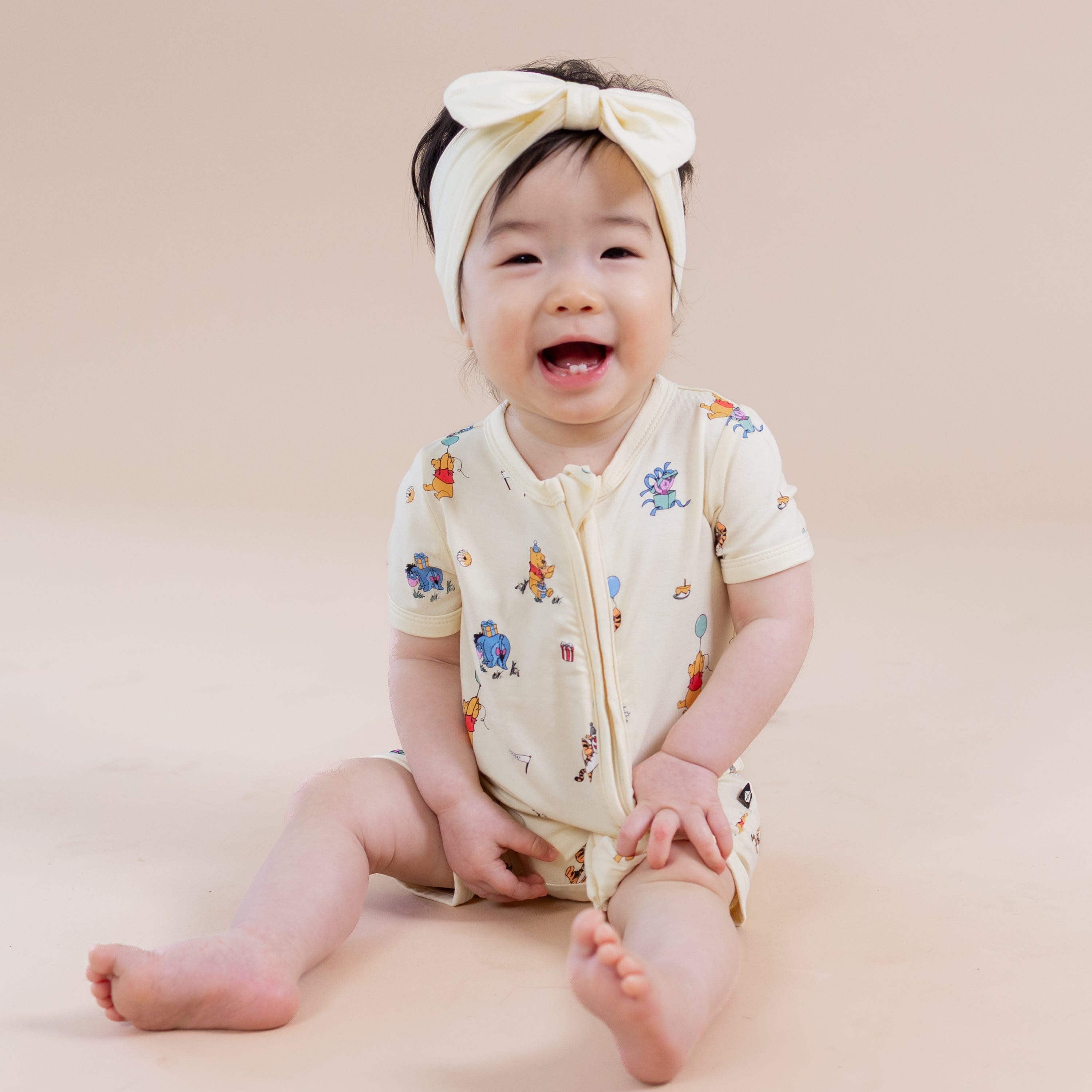 Young smiling girl sitting wearing the Zipper Shortall Romper in Winnie the Pooh Celebration