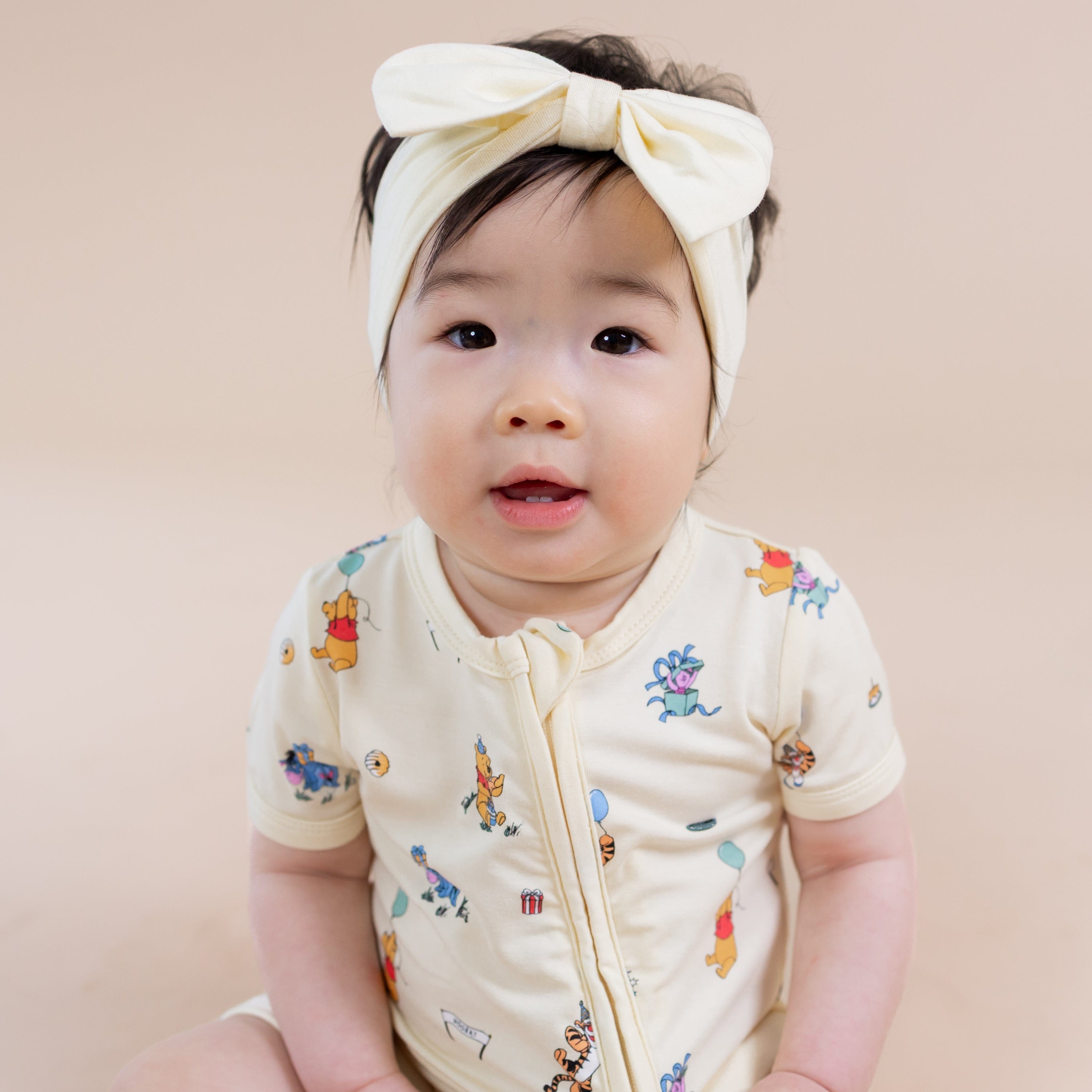 Close up of the Zipper Shortall Romper in Winnie the Pooh Celebration shown on a toddler wearing light yellow bow