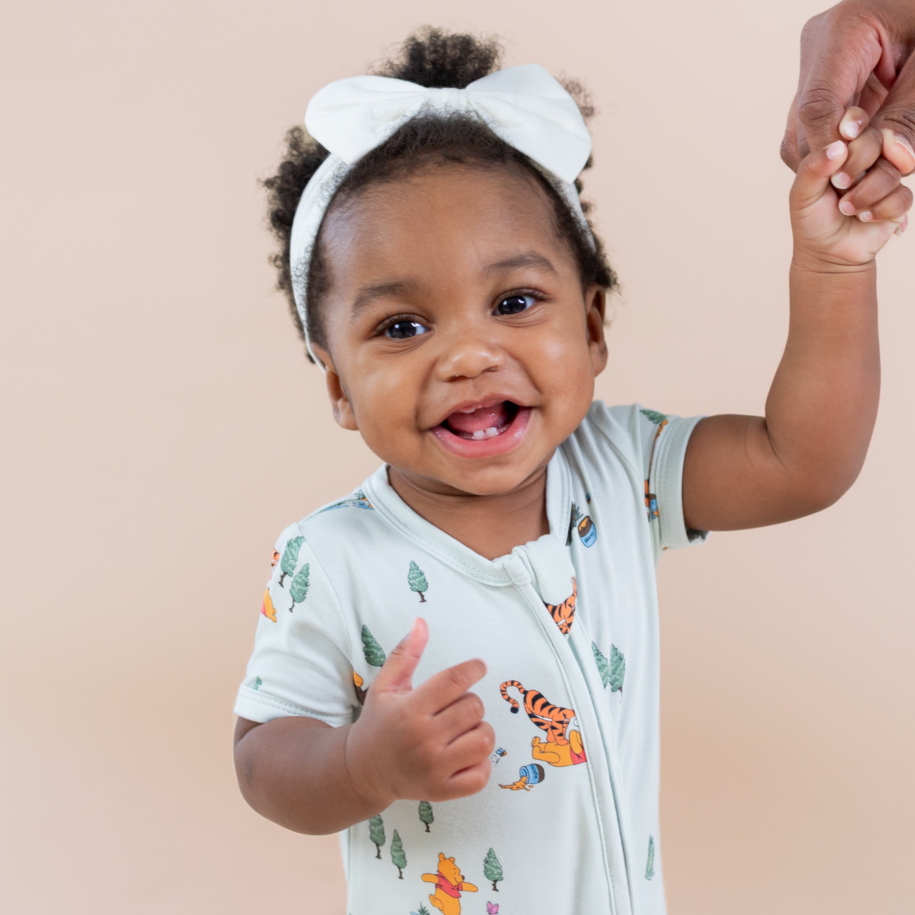 Close up of young toddler holding her mothers hand wearing the Zipper Shortall Romper in Winnie the Pooh Hundred Acre Wood paired with a white bow
