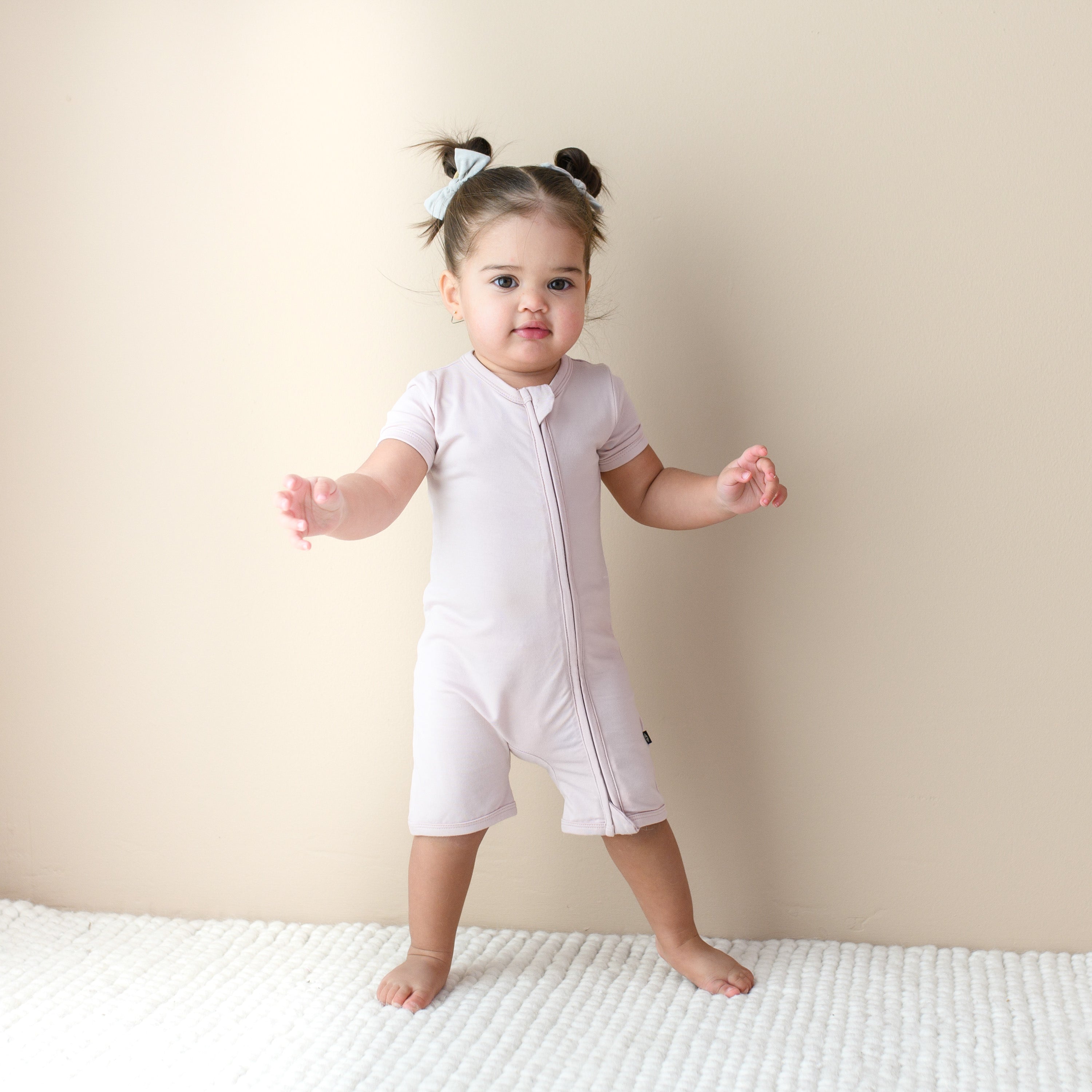 Toddler standing infront of beige wall while wearing dual Zipper Shortall Romper in Blush