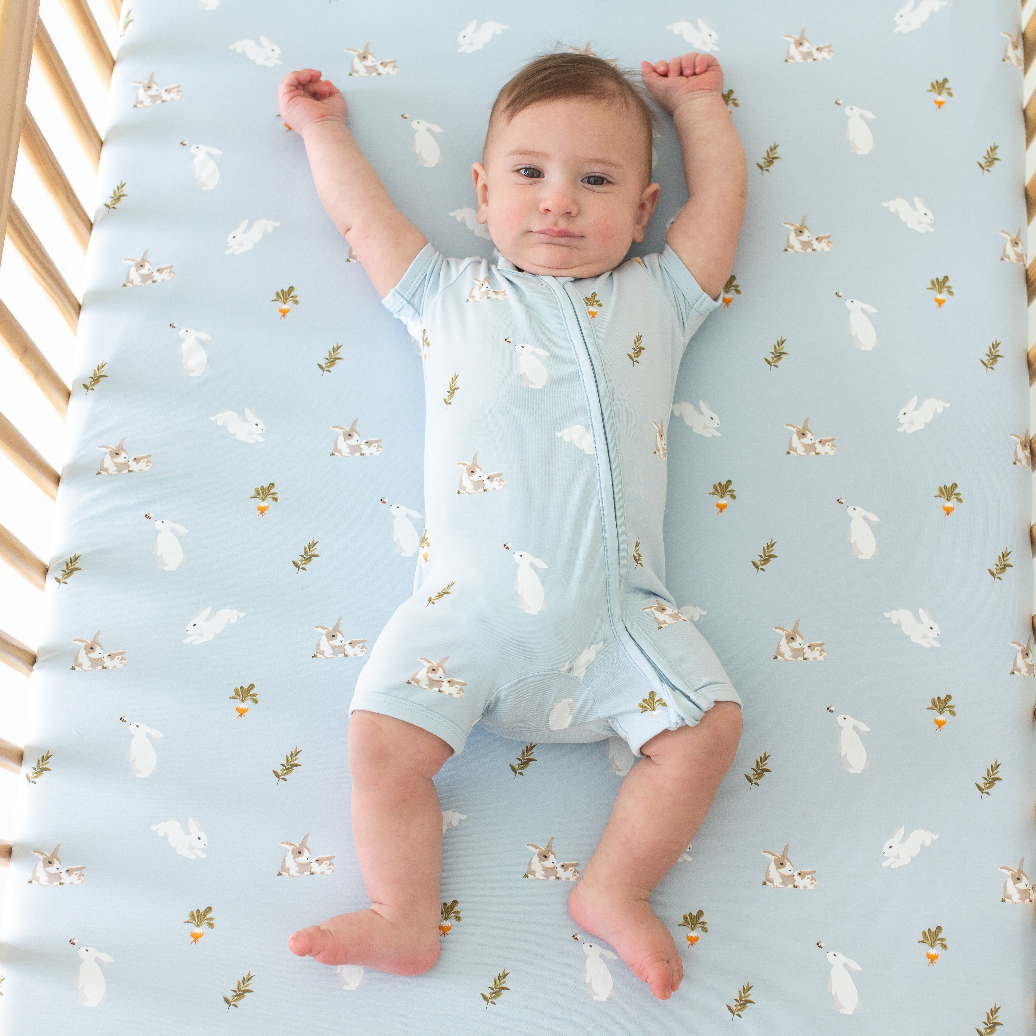 Infant wearing Kyte Baby Zippered Shortall Romper in Ice Rabbit and laying on matching Crib Sheet