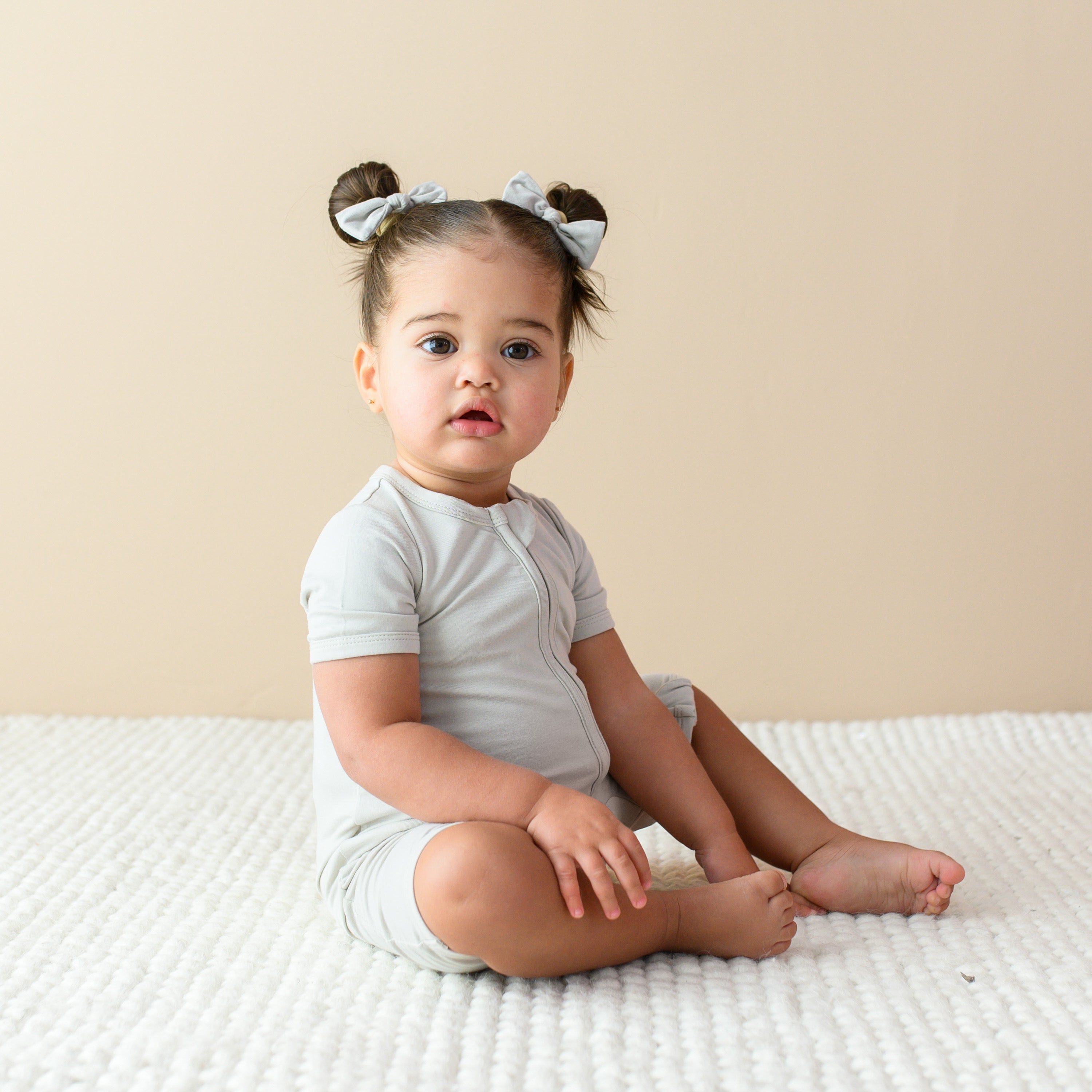 Toddler sitting on a cream colored blanket while wearing a Zippered Shortall Romper in Oat and matching bows in buns