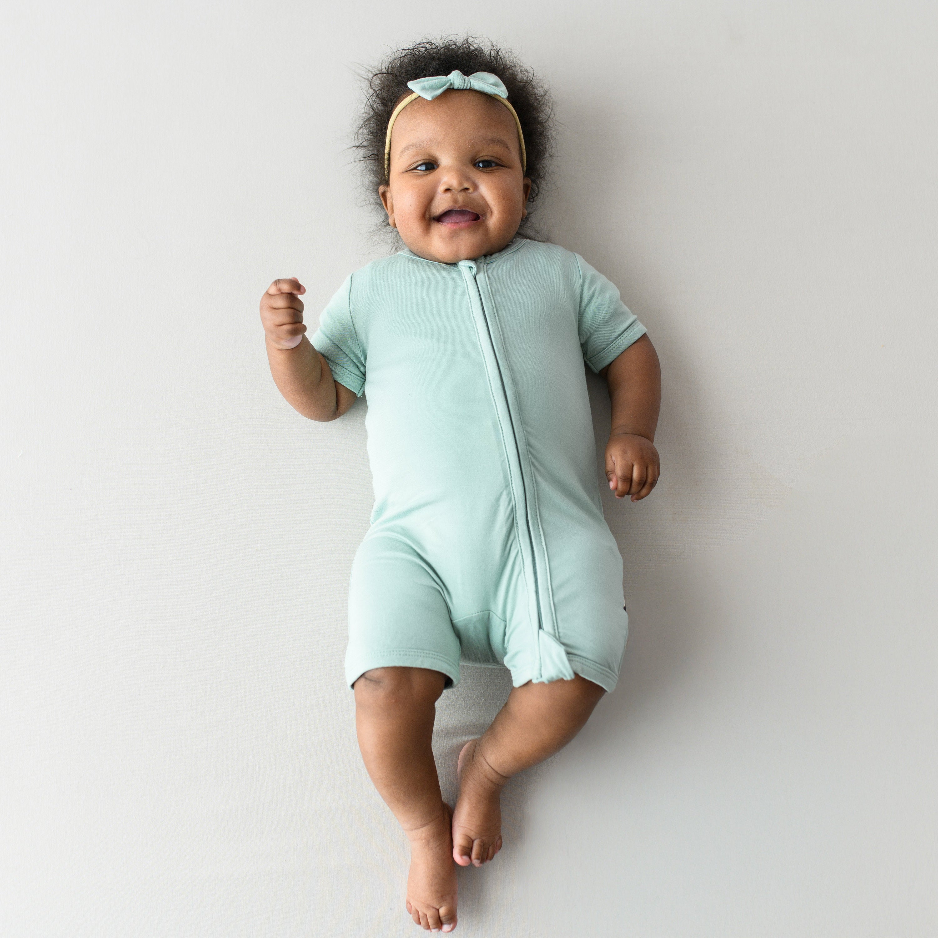 Baby laying on a oat background while wearing a Zippered Shortall Romper in Sage and matching bow