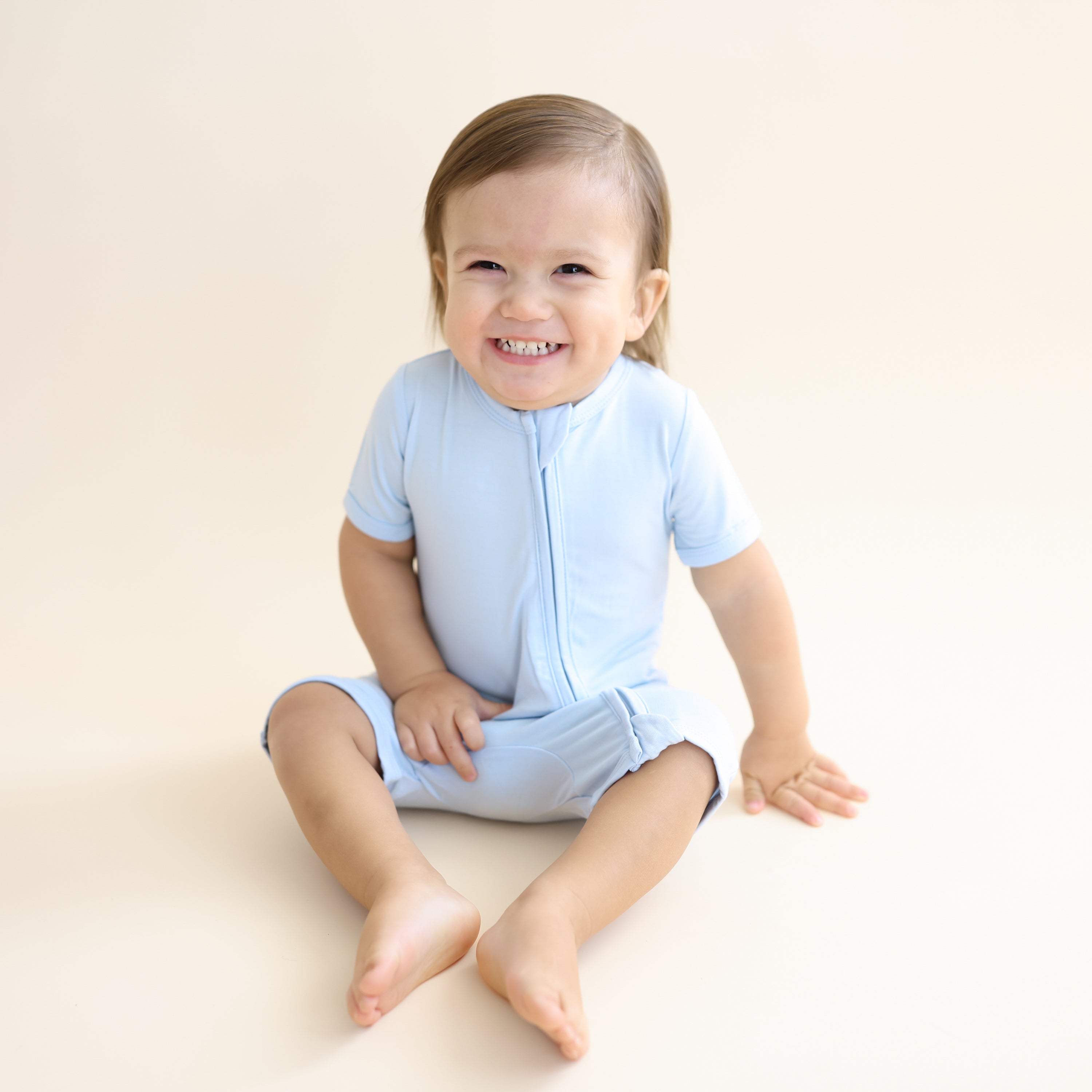 Young smiling boy sitting on the floor wearing the Zipper Shortall Romper in Breeze