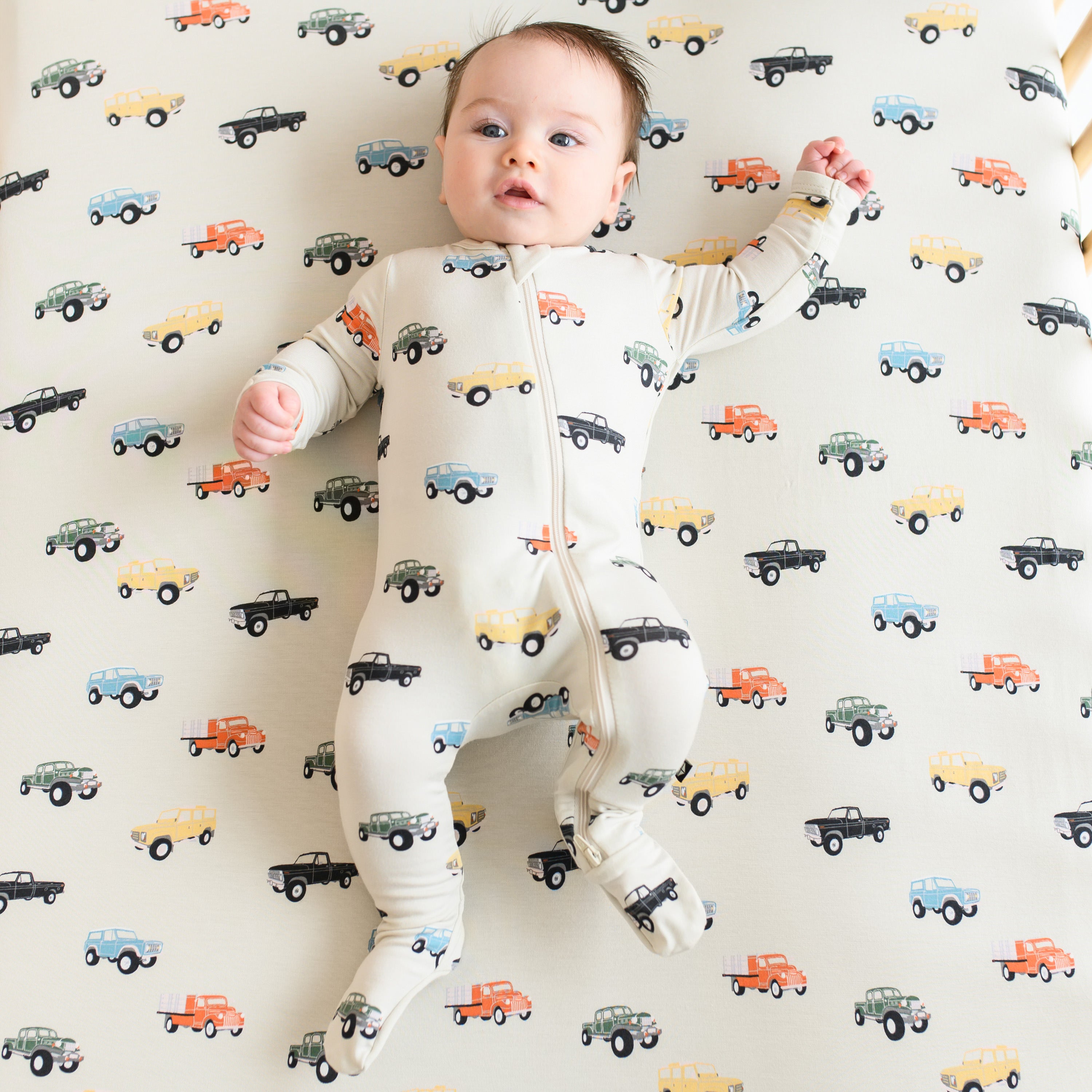 Infant wearing the Zippered Footie pajamas in Vintage Truck laying on a matching crib sheet