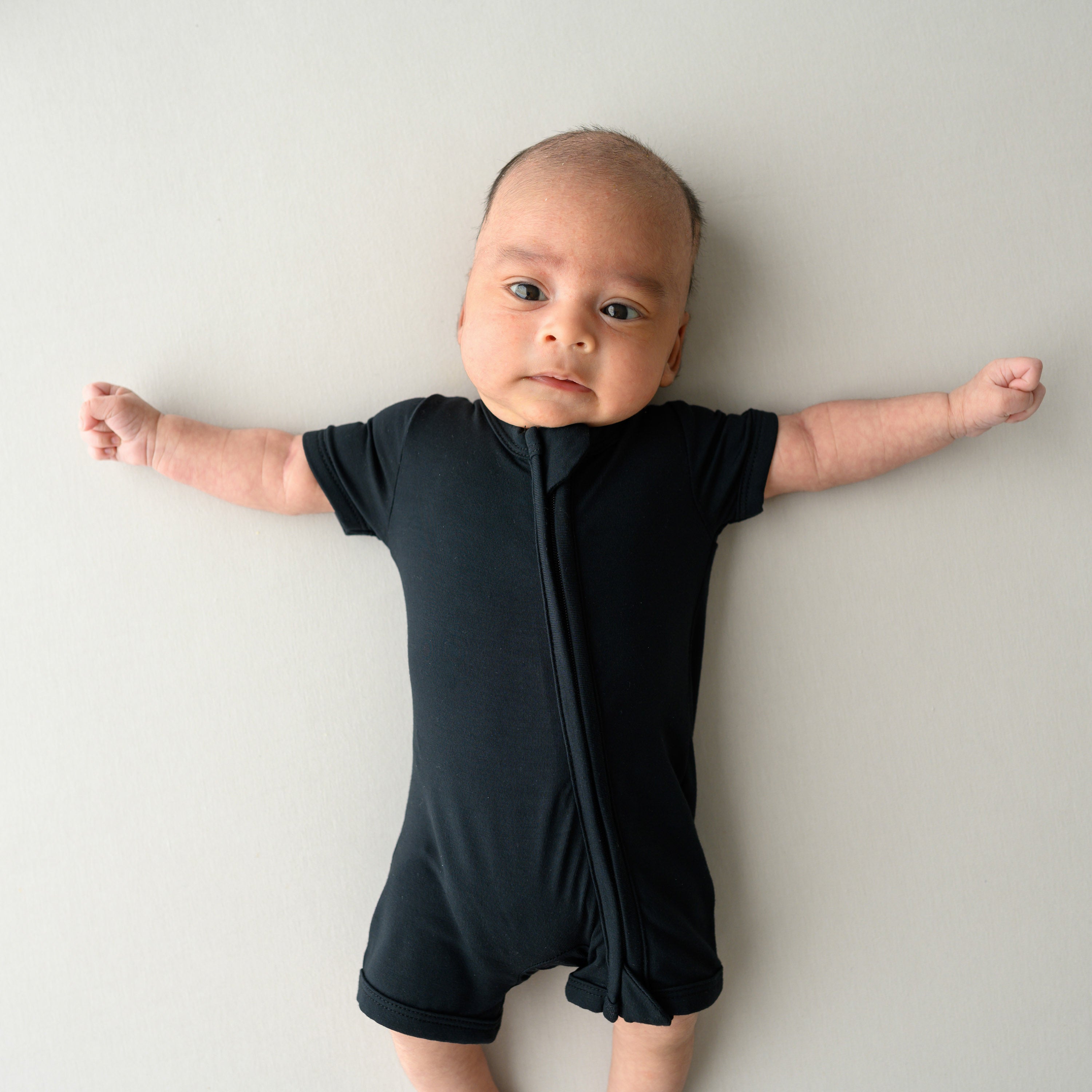 Infant laying on an oat surface while wearing a Zippered Shortall Romper in Midnight
