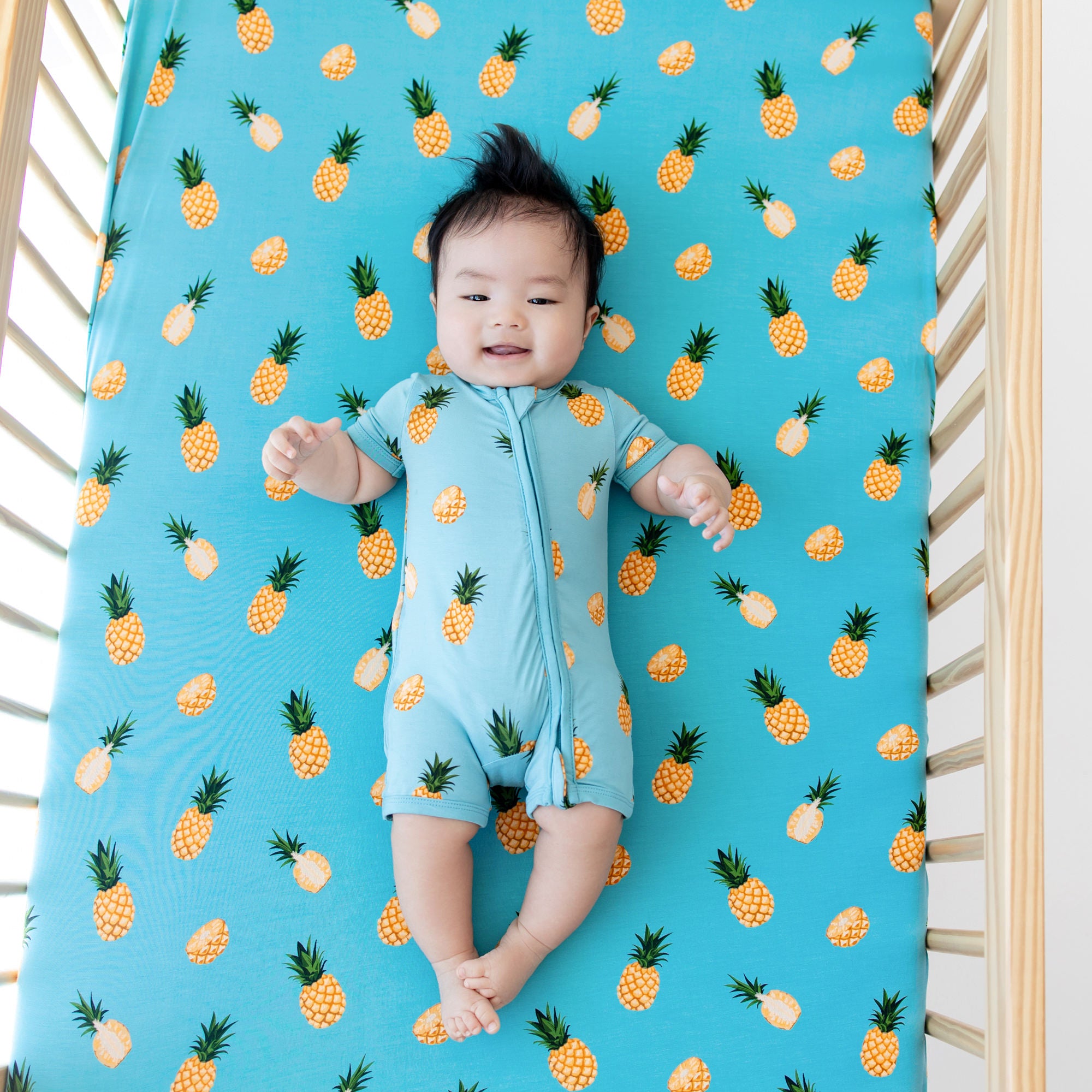 Infant wearing Kyte Baby Zippered Shortall Romper in Pineapple laying on matching Crib Sheet