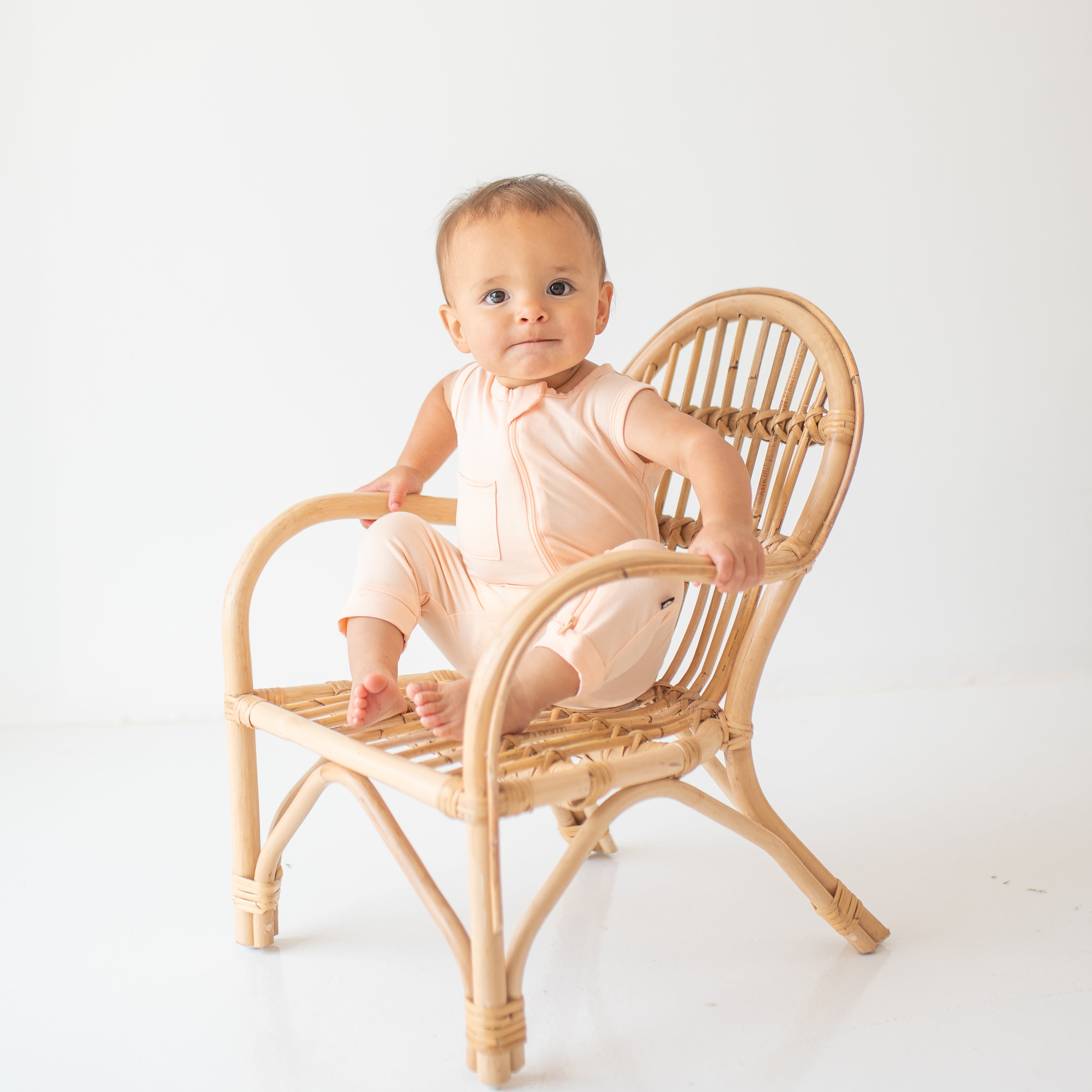 Infant model sitting on a wicker chair wearing the Zippered Sleeveless Romper in Canyon