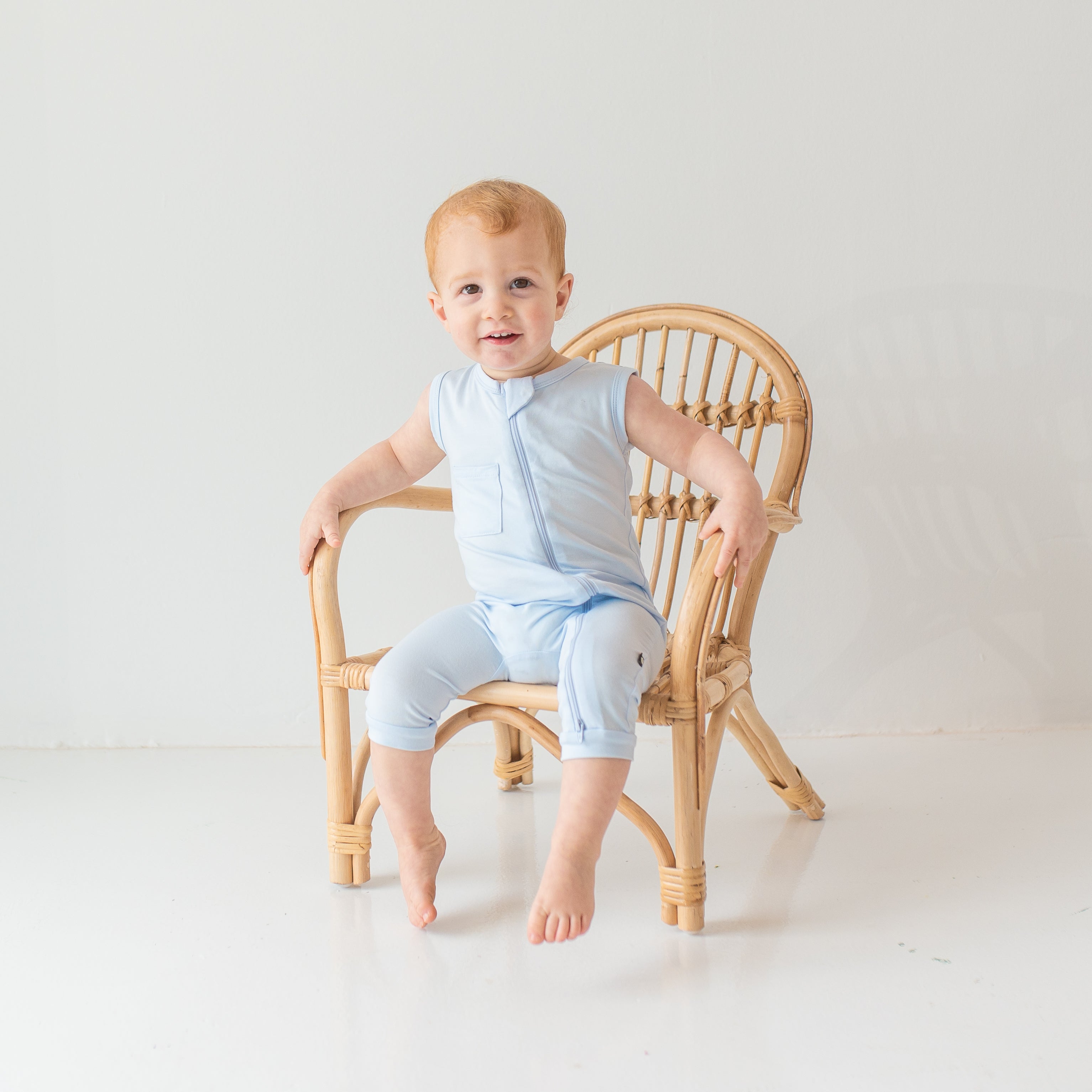 Young toddler model sitting on a wicker chair wearing the Zippered Sleeveless Romper in Mist