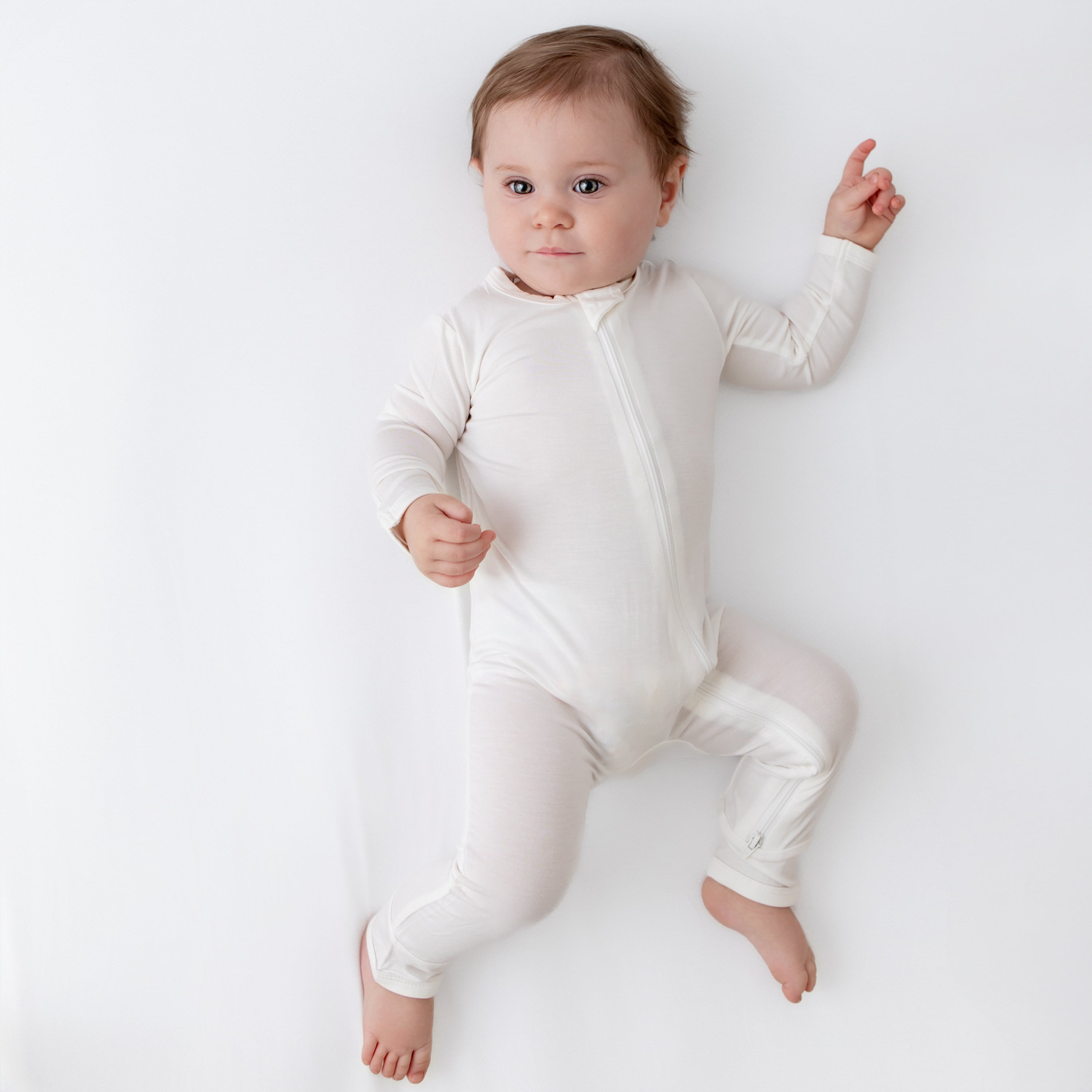 Baby laying on a Cloud surface with one hand up and wearing a Zippered Romper in Cloud