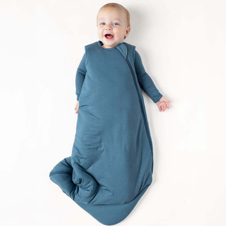 Smiling infant laying down wearing the Sleep Bag in Atlantic 2.5 with a matching long sleeve romper on underneath
