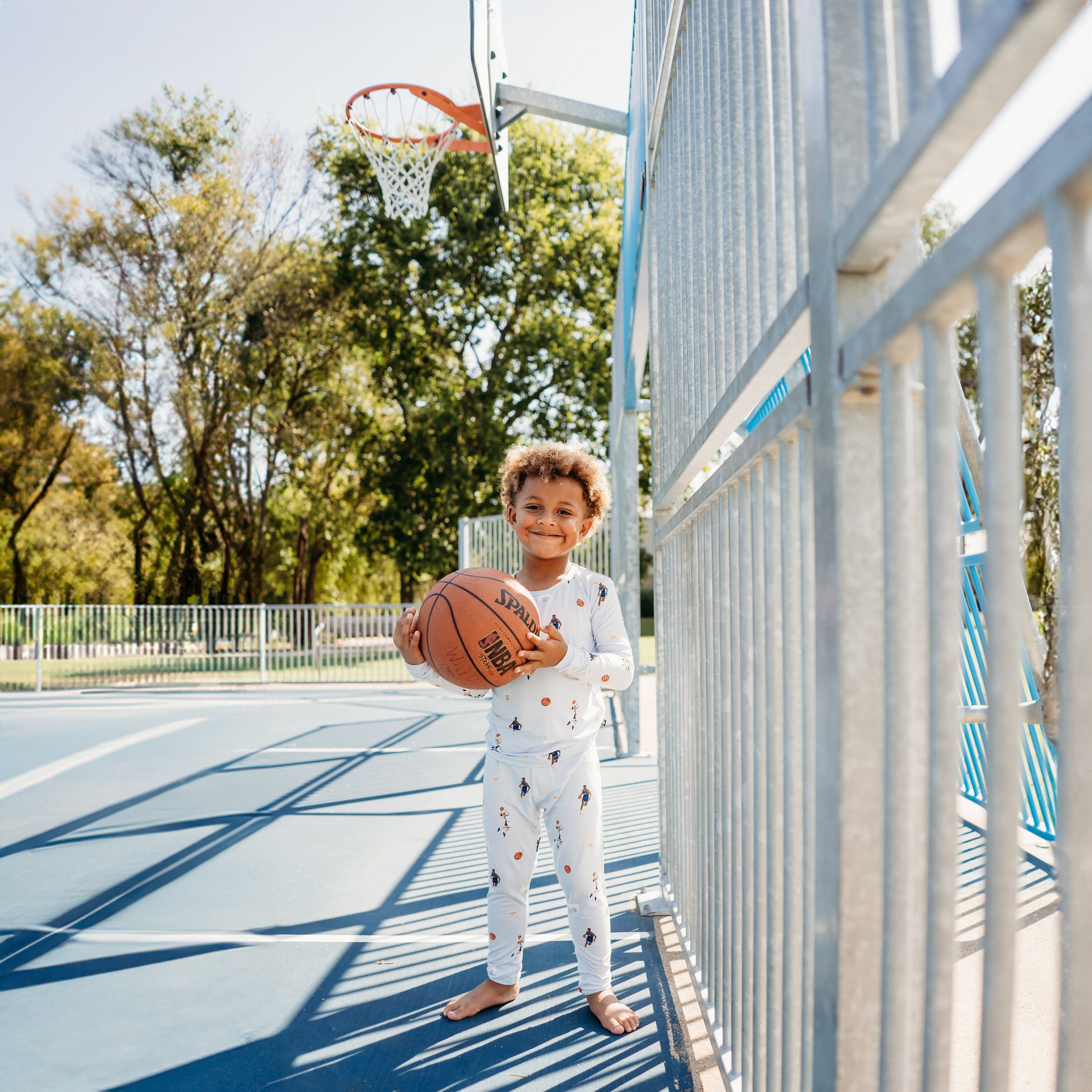 Young boy smiling holding a basketball standing on a basketball court wearing the Long Sleeve Pajamas in Basketball