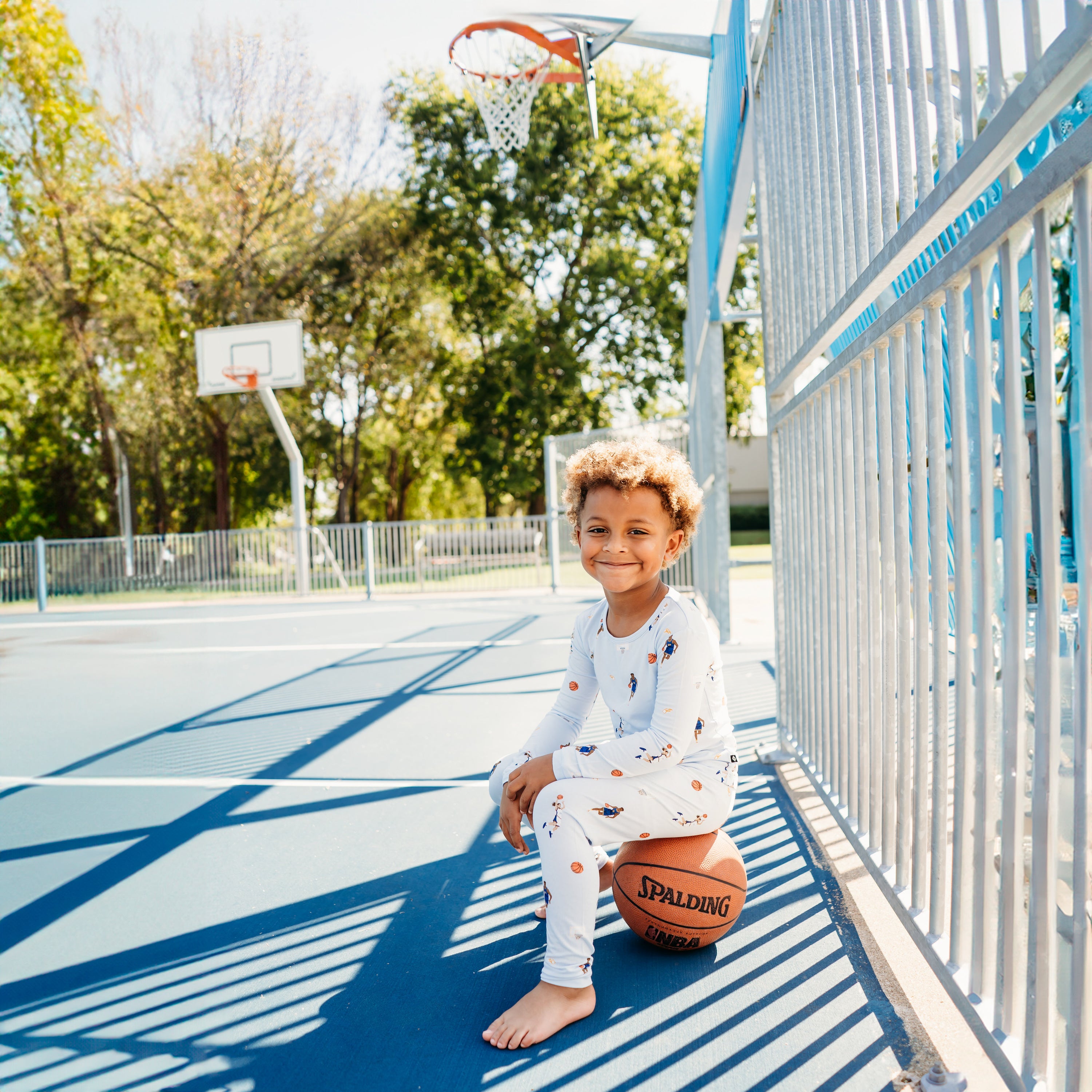 Smiling boy sitting on a basketball on a basketball court wearing the Long Sleeve Pajamas in Basketball