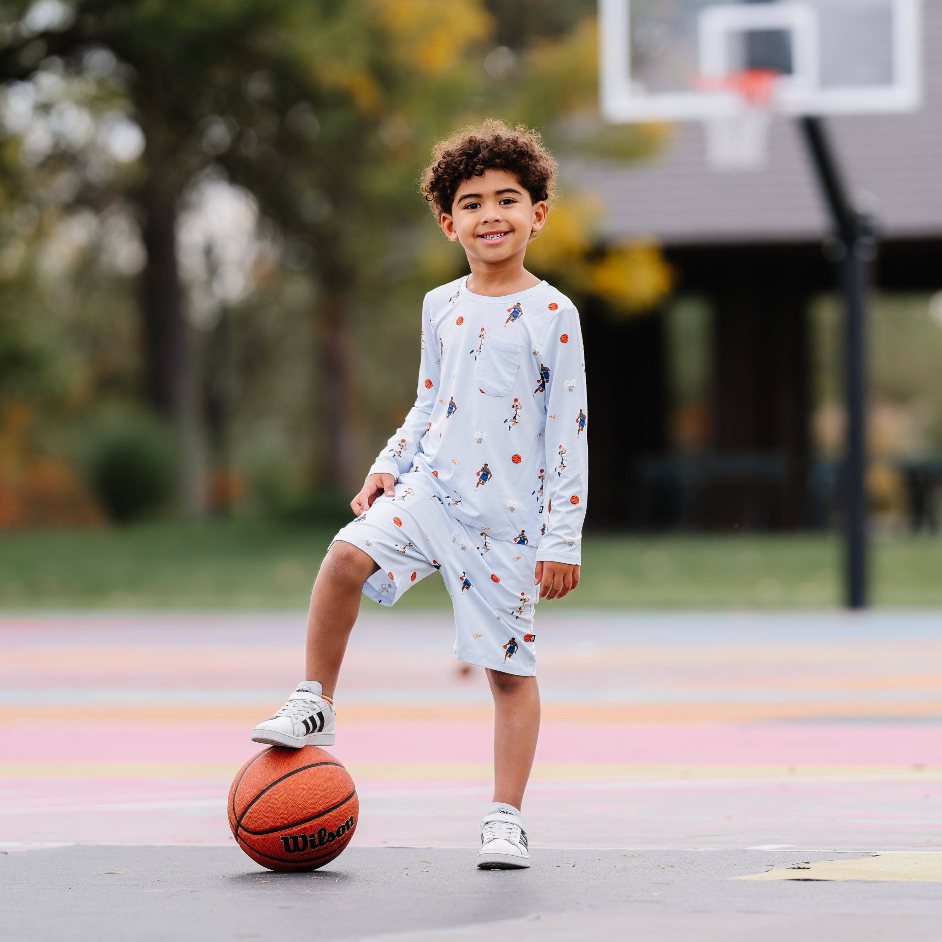 Young boy standing on a basketball court with one foot on a basketball wearing the Long Sleeve Toddler Crew Neck Tee in Basketball and matching drawstring shorts