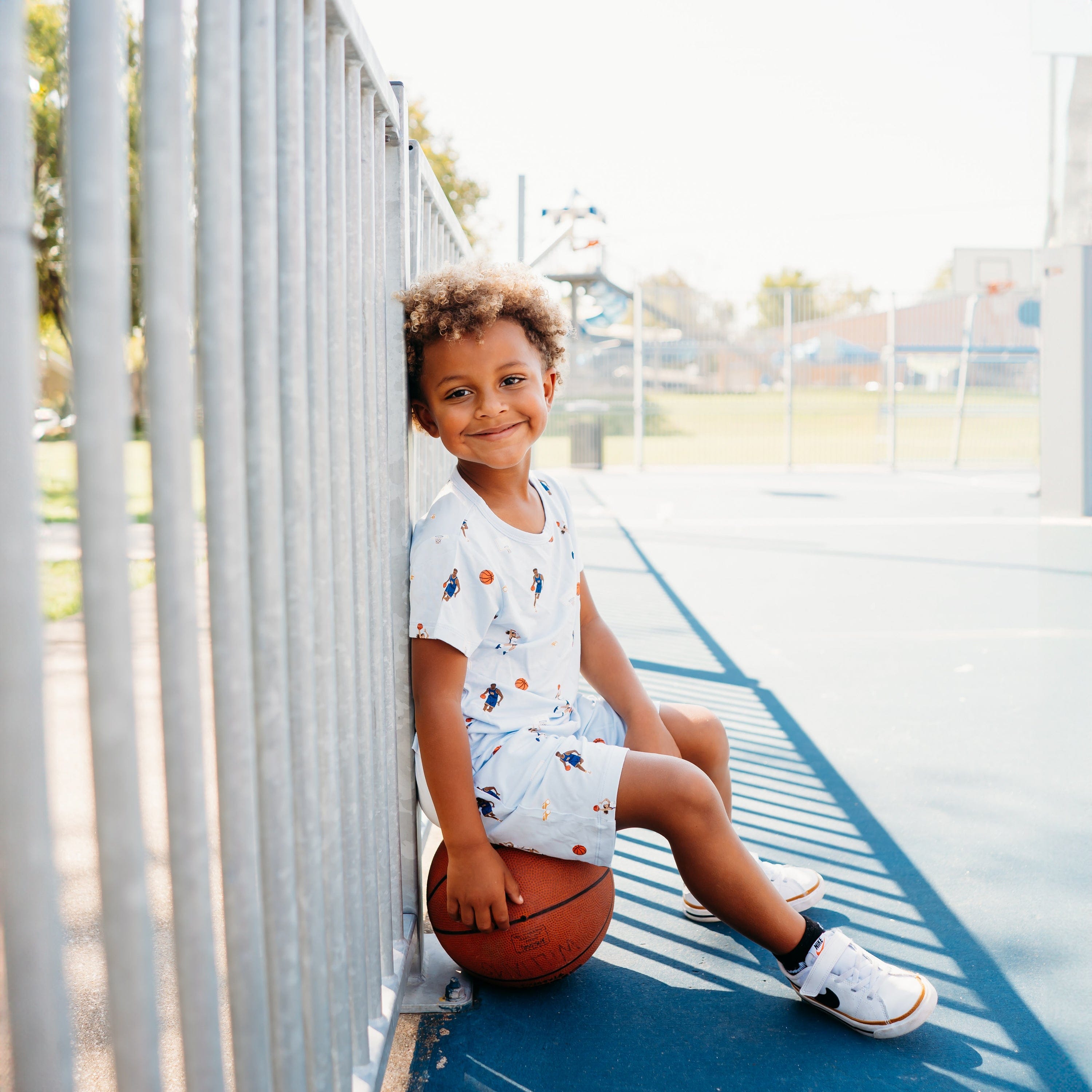Young boy sitting on a basketball leaning against a fence wearing the Toddler Basic Tee in Basketball and matching drawstring shorts