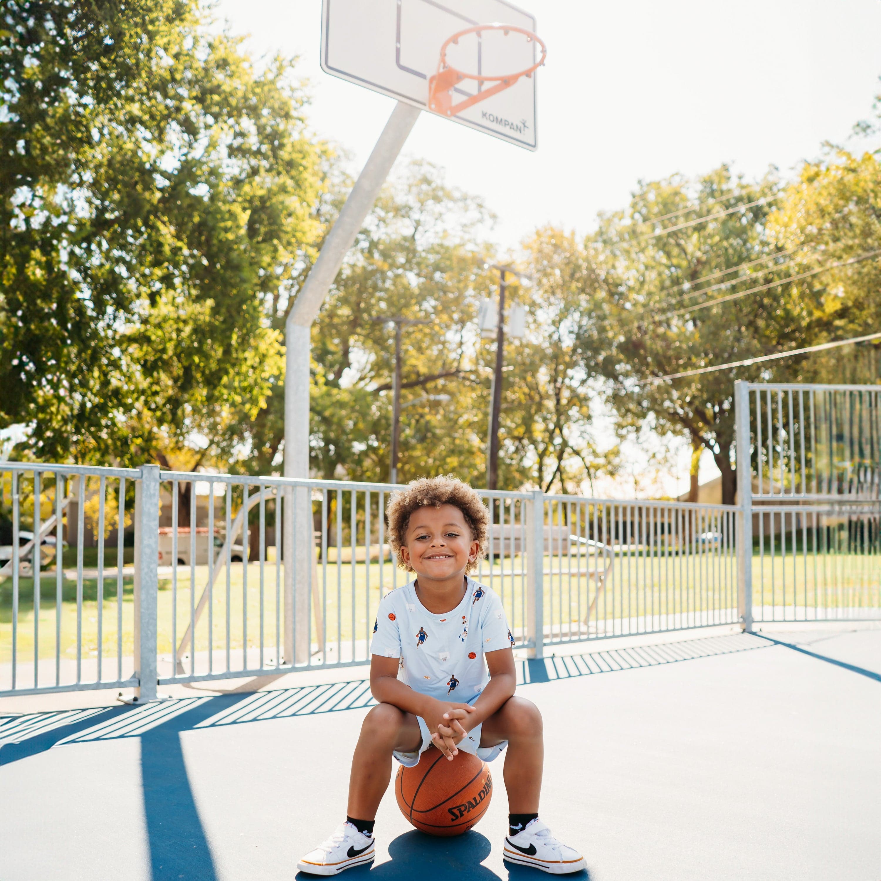 Young boy sitting on a basketball on a basketball court wearing the Toddler Basic Tee in Basketball