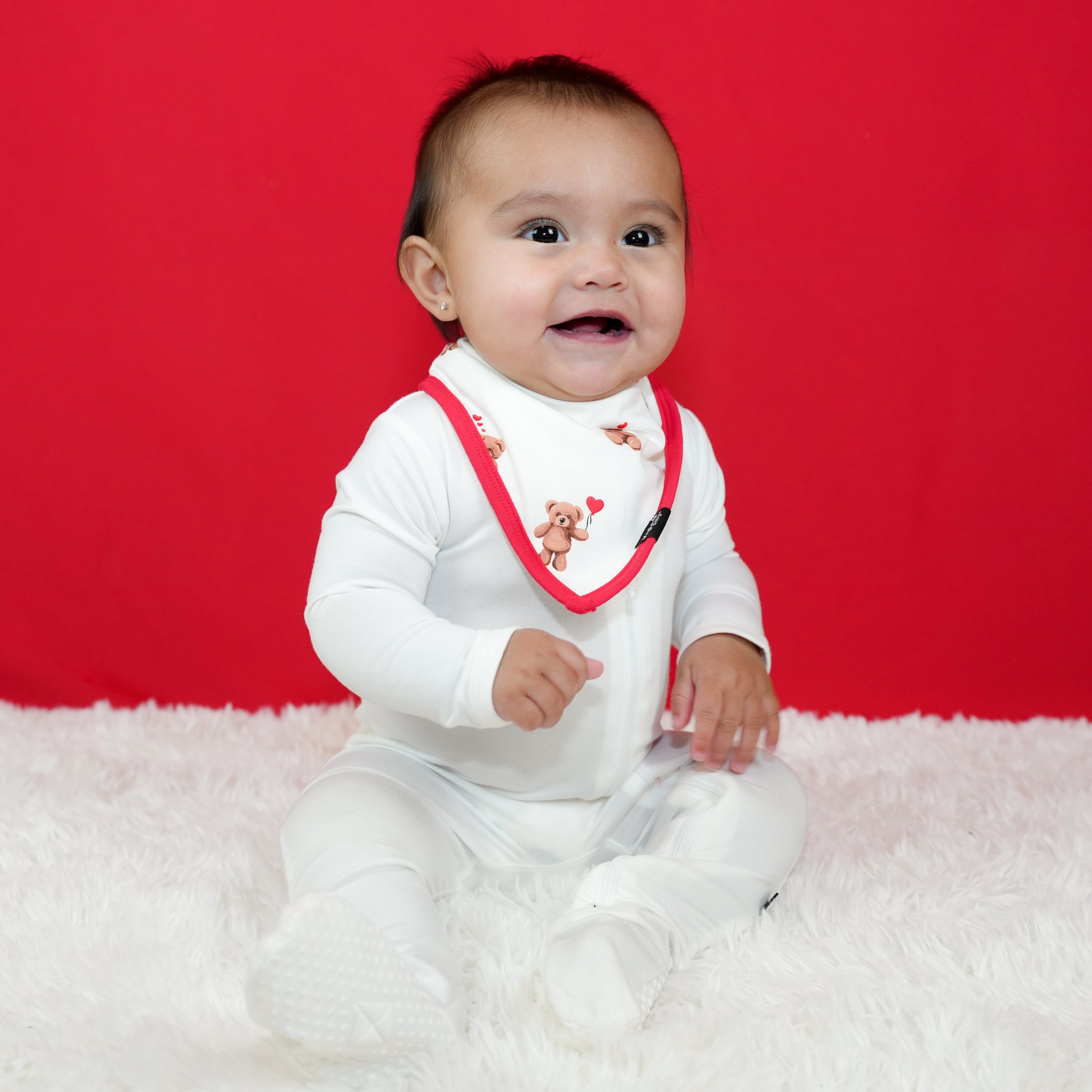Infant sitting on a white plush carpet in front of a red background wearing a white footie with a Bib in Bear Hearts