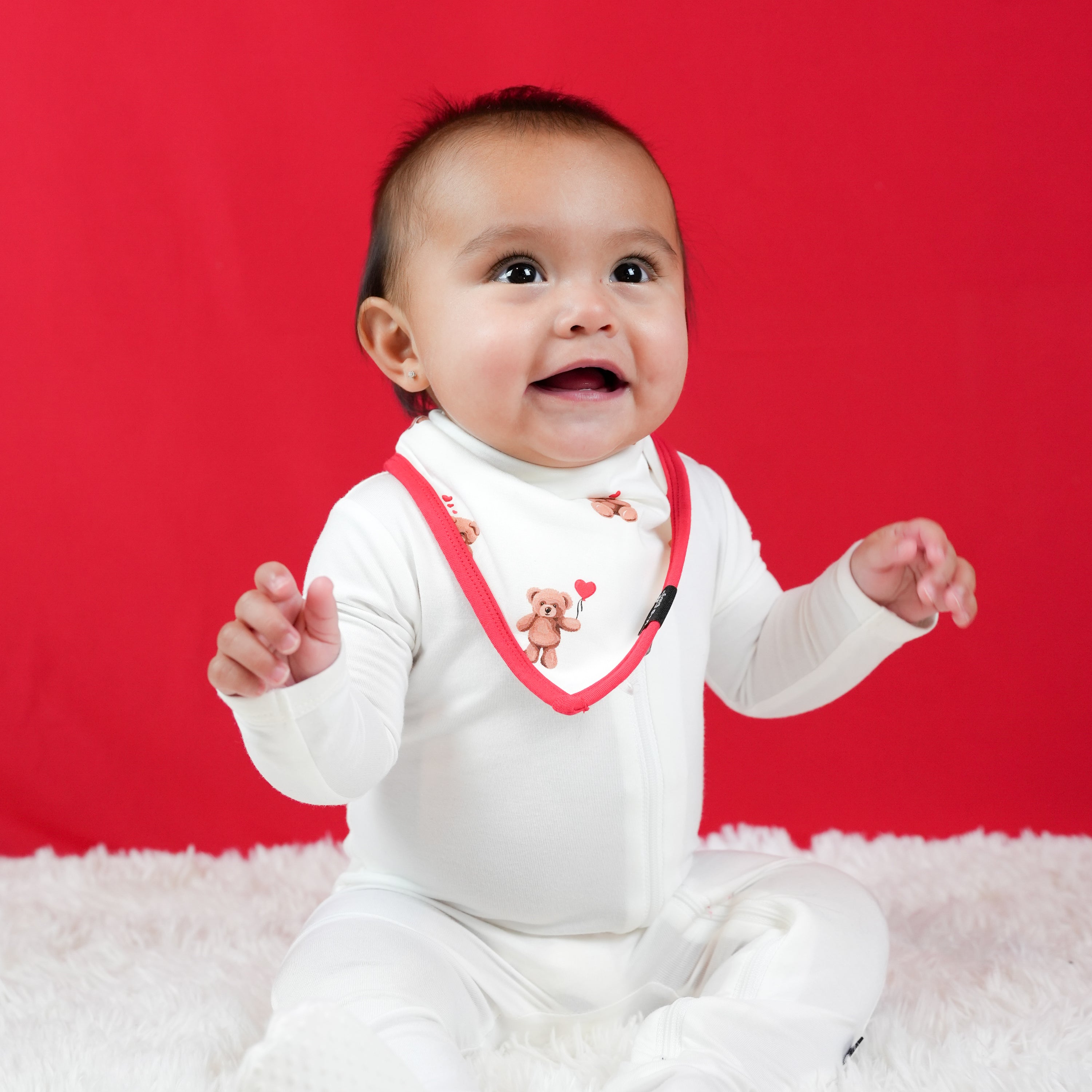 Young infant sitting on a white plush blanket in front of a red background wearing a white footie with the absorbant Bib in Bear Hearts