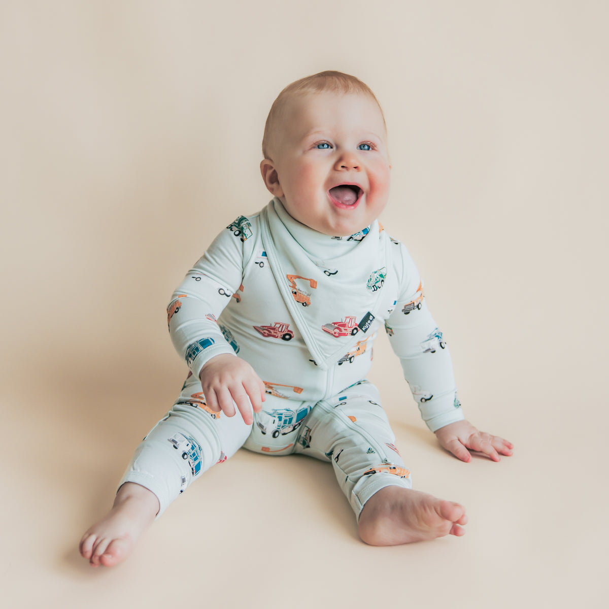 Smiling toddler sitting on the floor wearing the Bib in City Vehicles with matching zippered romper