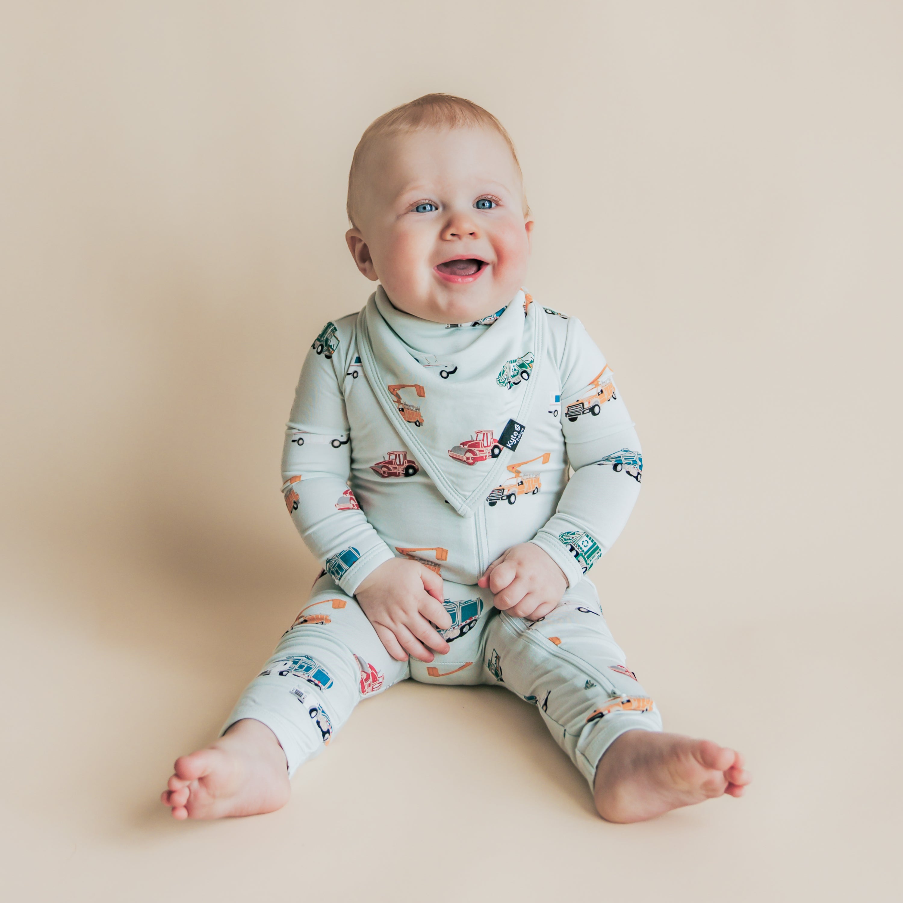 Toddler sitting on a light beige neutral surface wearing the soft and absorbent Bib in City Vehicles with a matching zippered romper