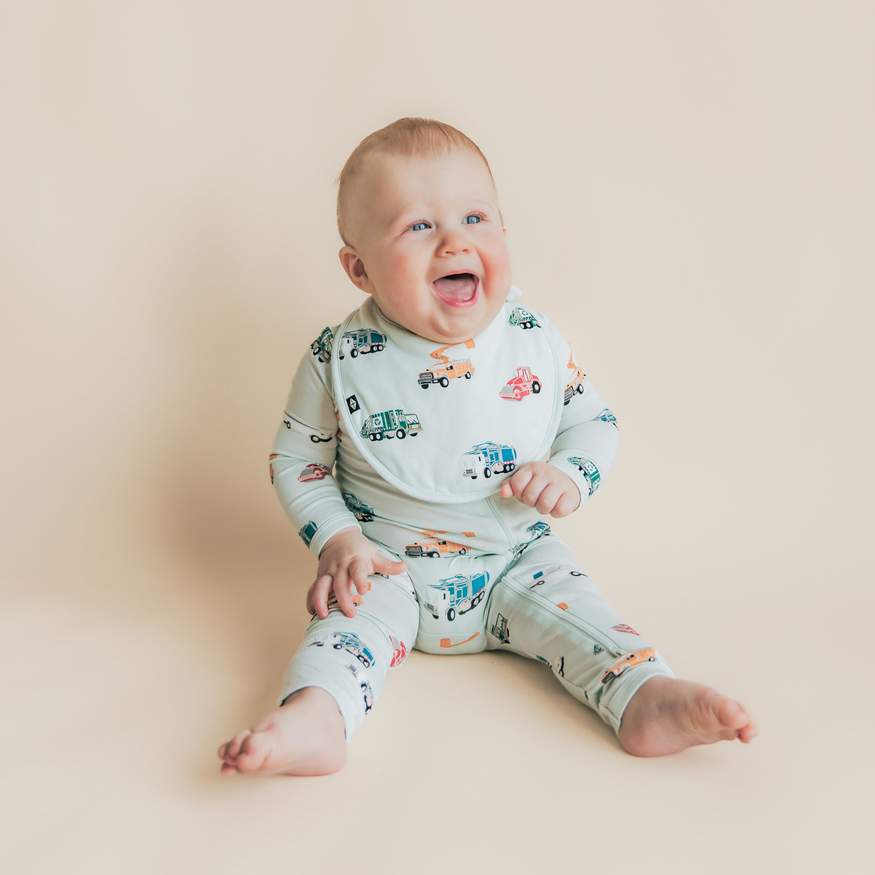 Smiling toddler sitting on the floor wearing the Cushy Bib in City Vehicles with matching zippered romper