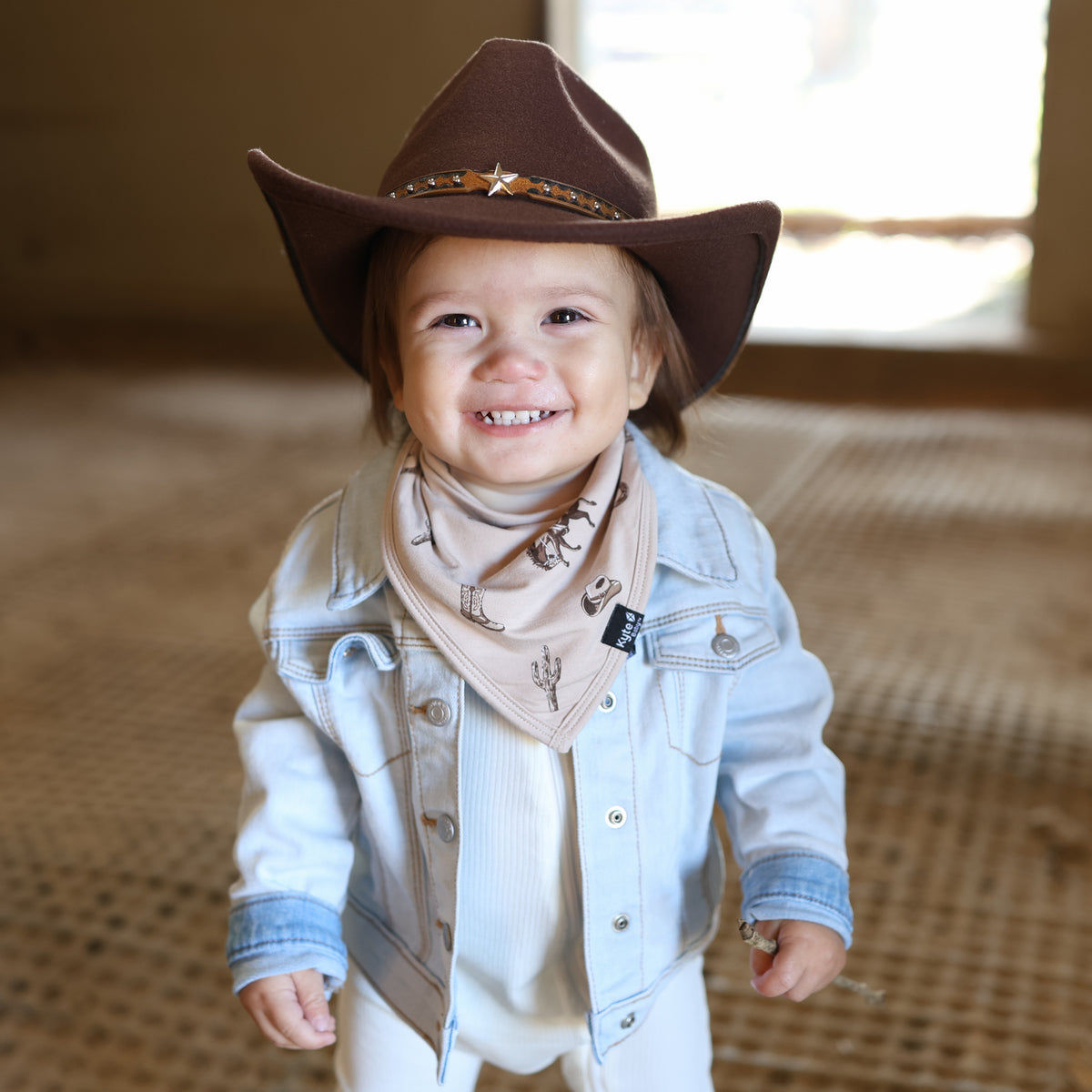 Smiling toddler wearing the absorbent Bib in Classic Cowboy paired with a brown western hat and jean jacket