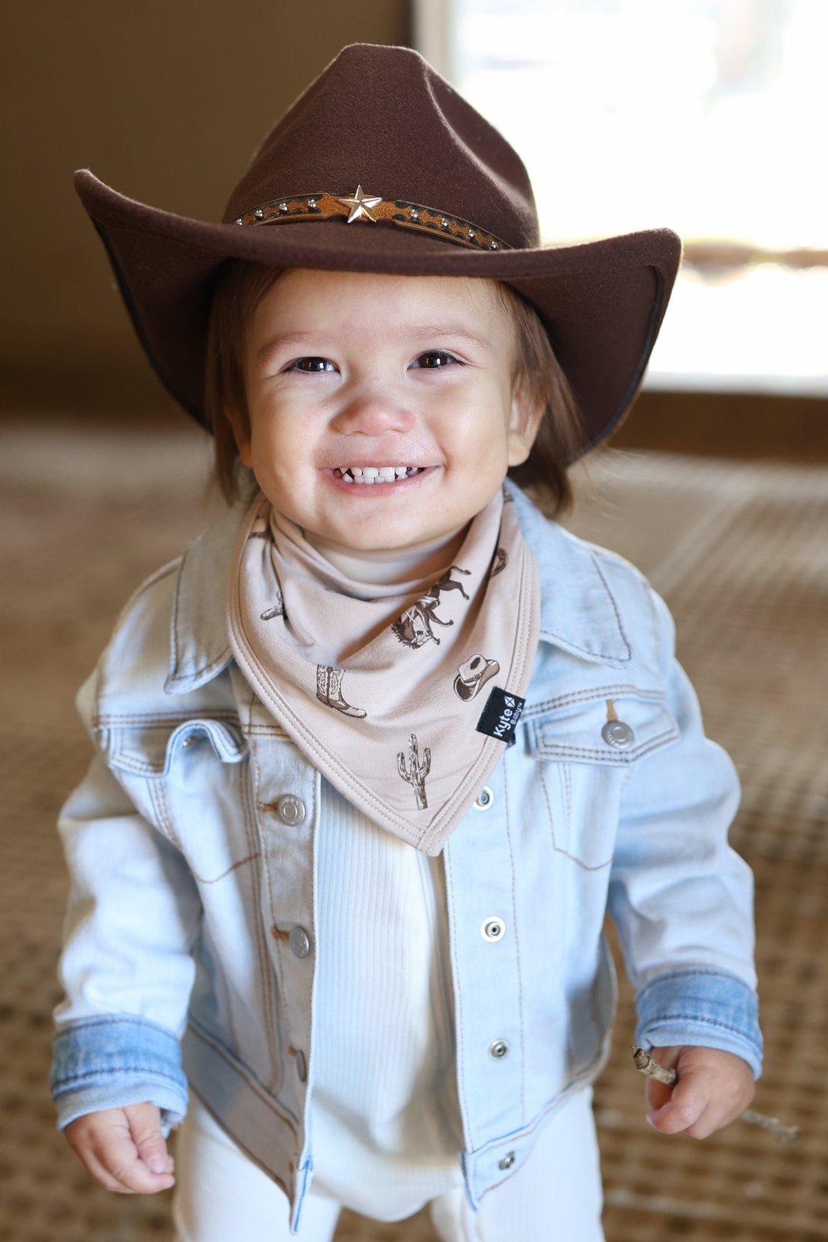 Smiling toddler wearing the absorbent Bib in Classic Cowboy paired with a brown western hat and jean jacket