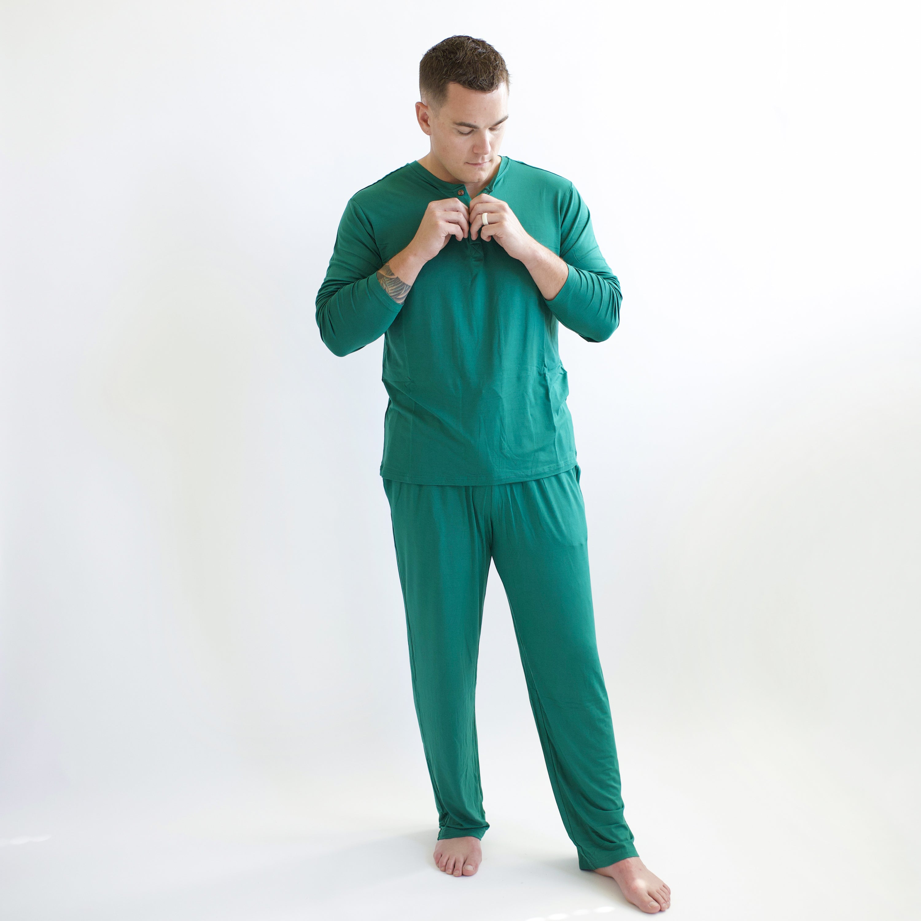 Male model standing in front of a white backdrop wearing the Men's Lounge Pants in Emerald and buttoning up the matching Emerald Men's Henley Top