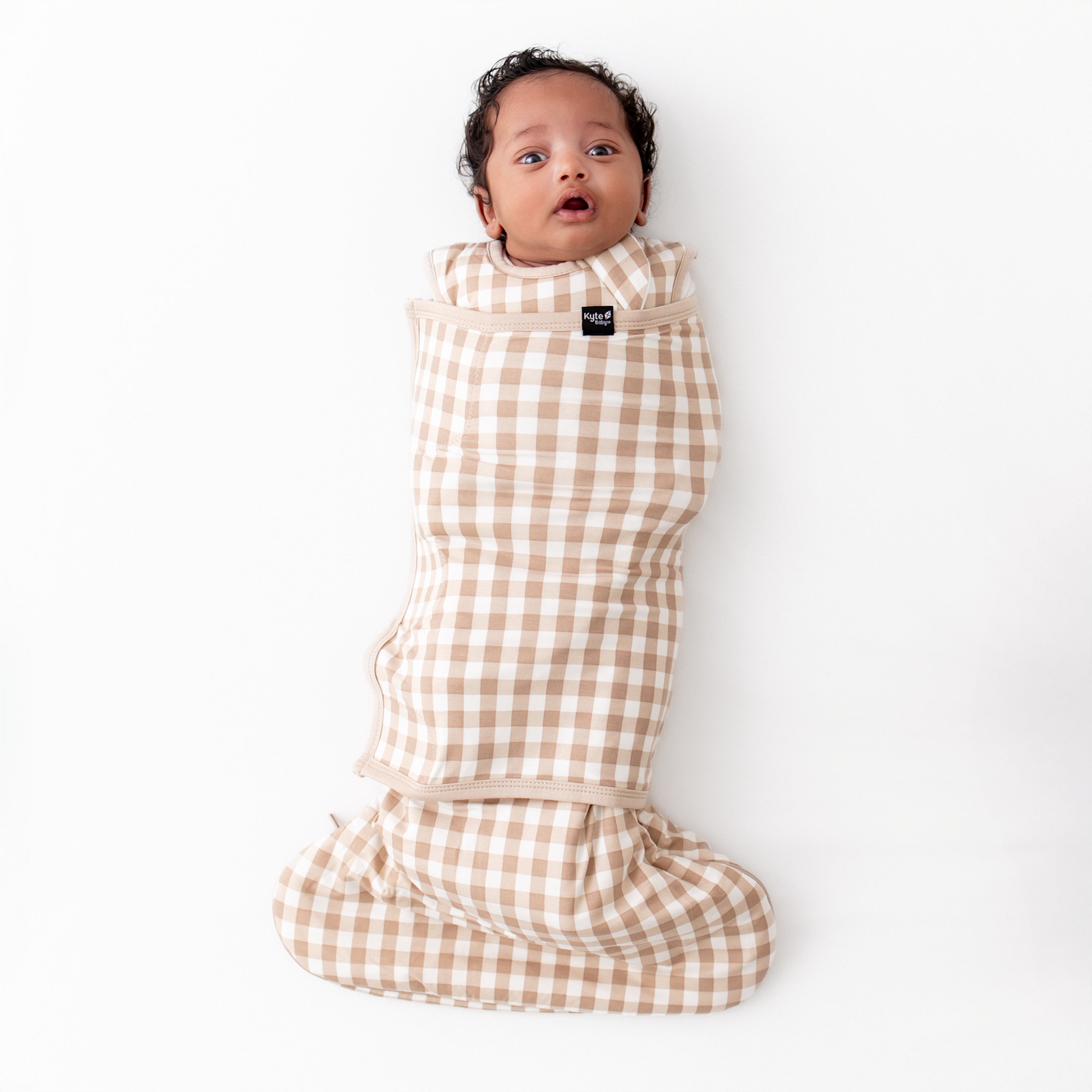 Infant laying down wearing the Sleep Bag Swaddler in Gingham Bisque 1.0