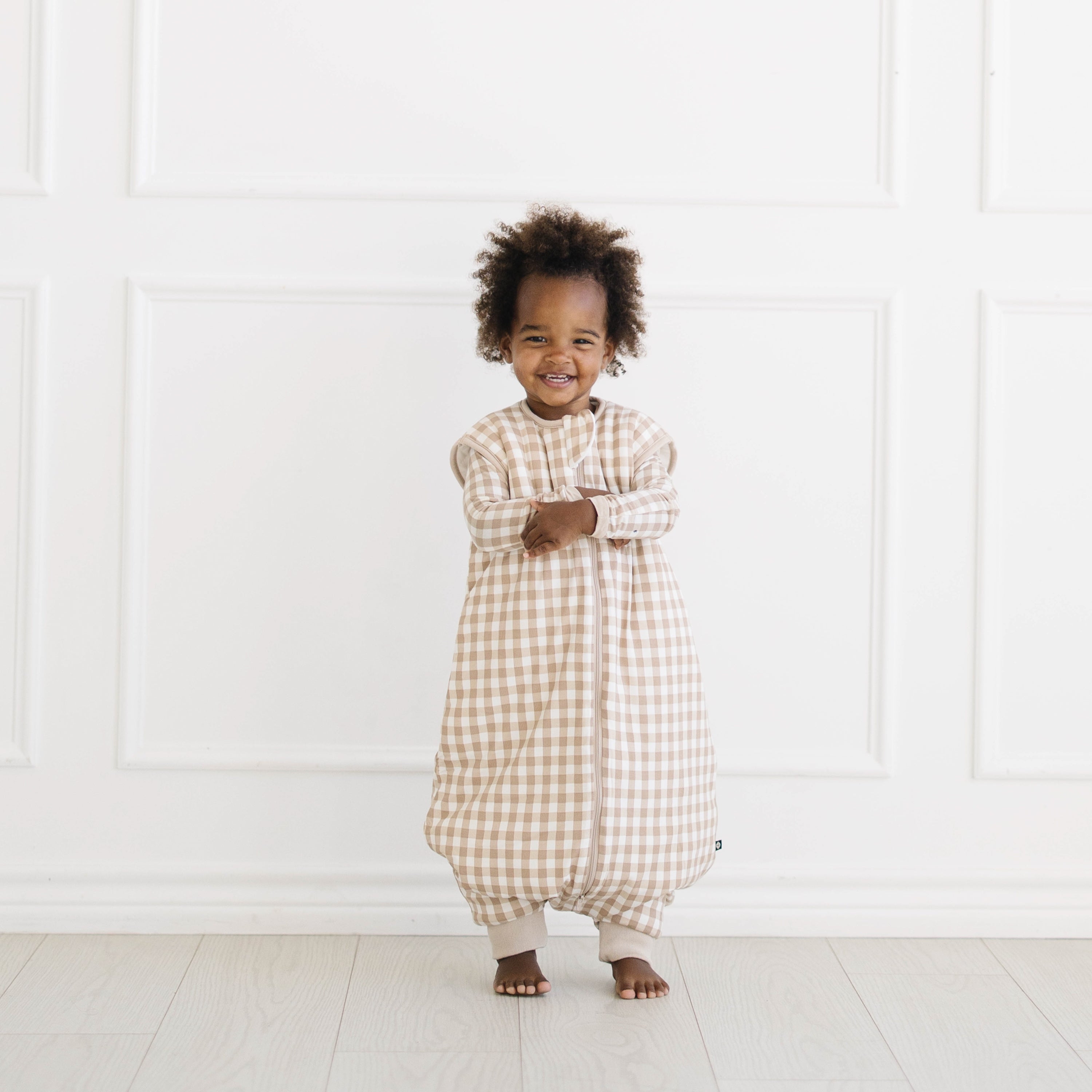 Smiling toddler standing wearing the Sleep Bag Walker in Gingham Bisque 2.5 with matching long sleeve toddler pajamas on underneath