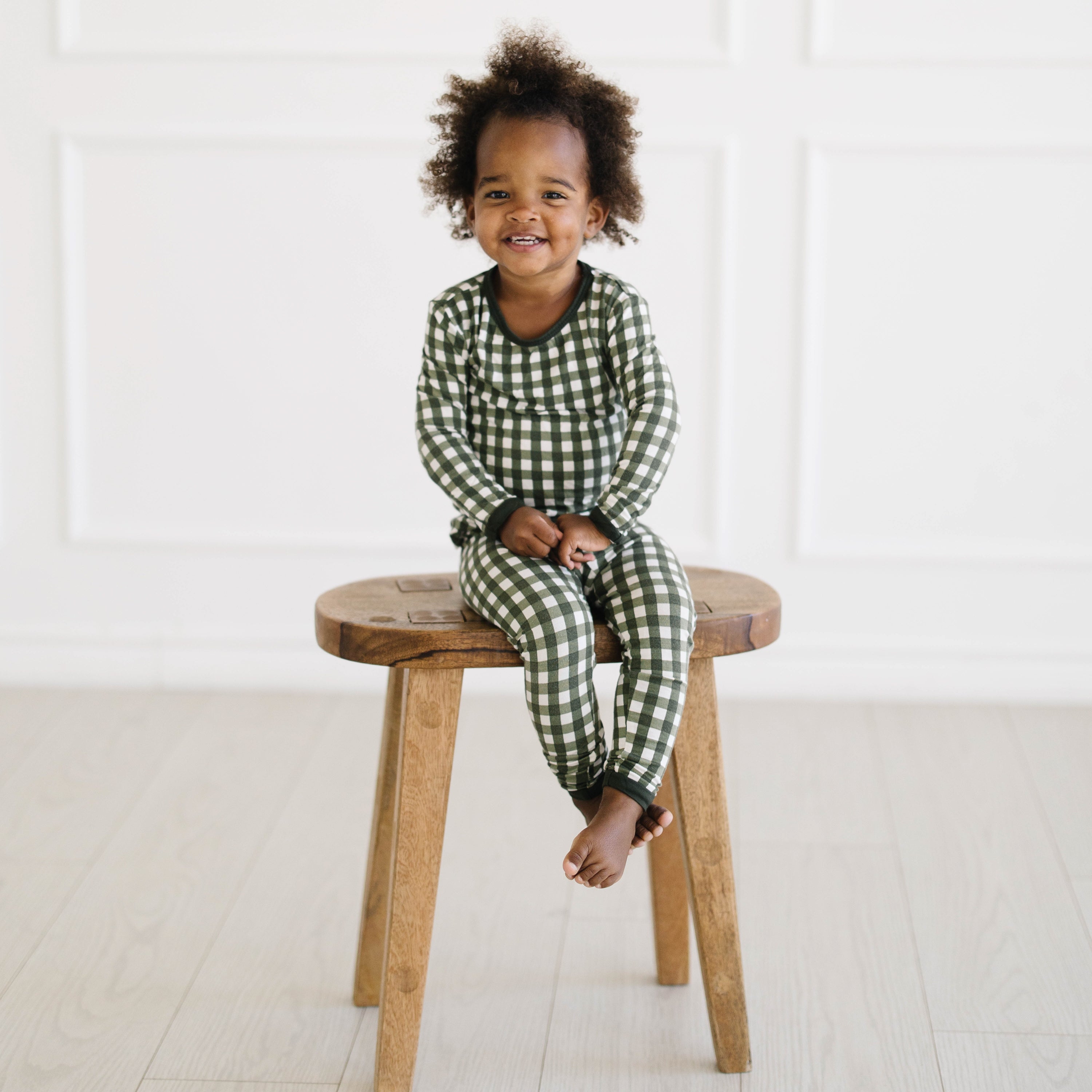 Young toddler sitting on a wooden stool wearing the Long Sleeve Pajamas in Gingham Fir