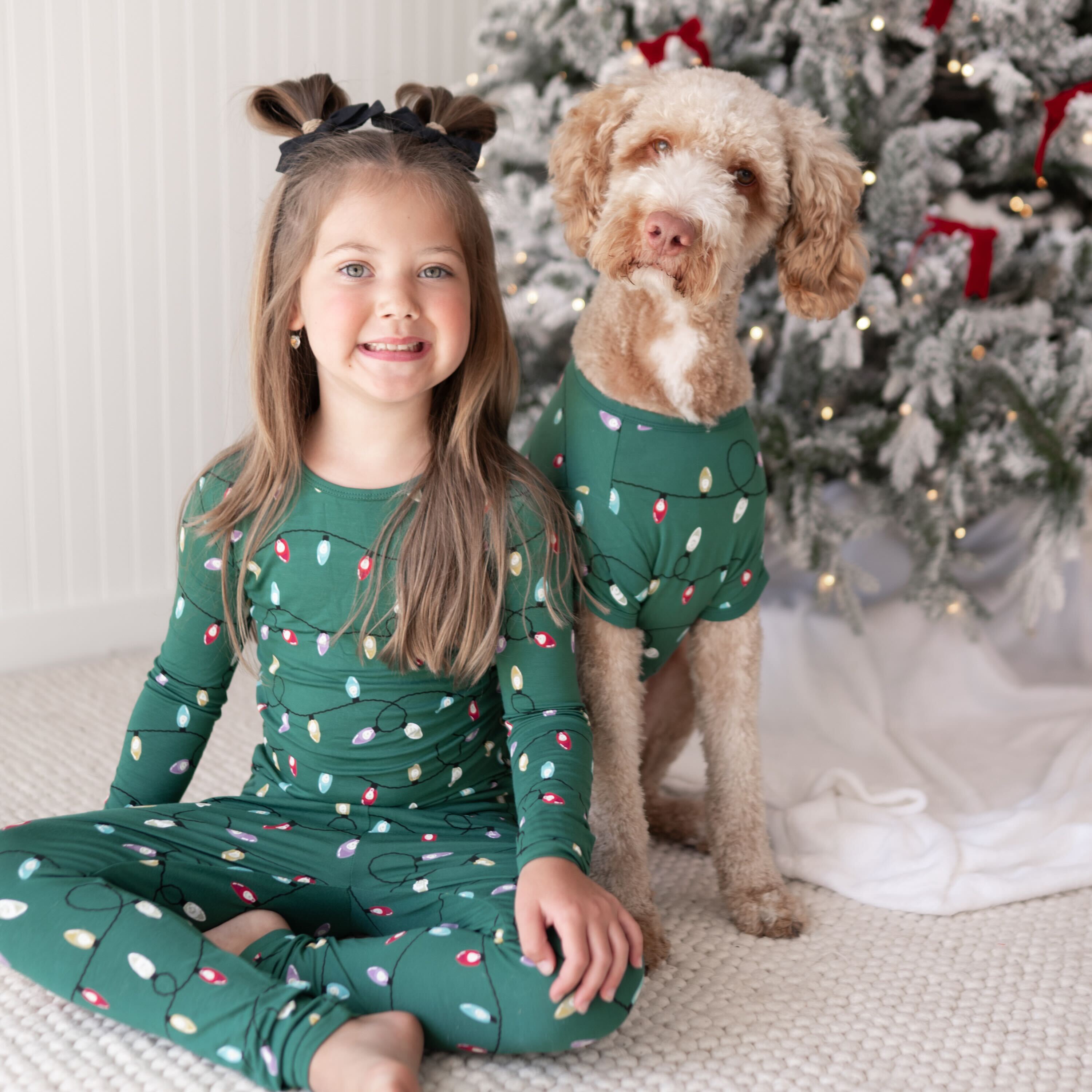 Young smiling girl wearing the Long Sleeve Pajamas in Merry and Bright sitting beside her dog who is wearing a matching dog tee both beside a decorated Christmas tree