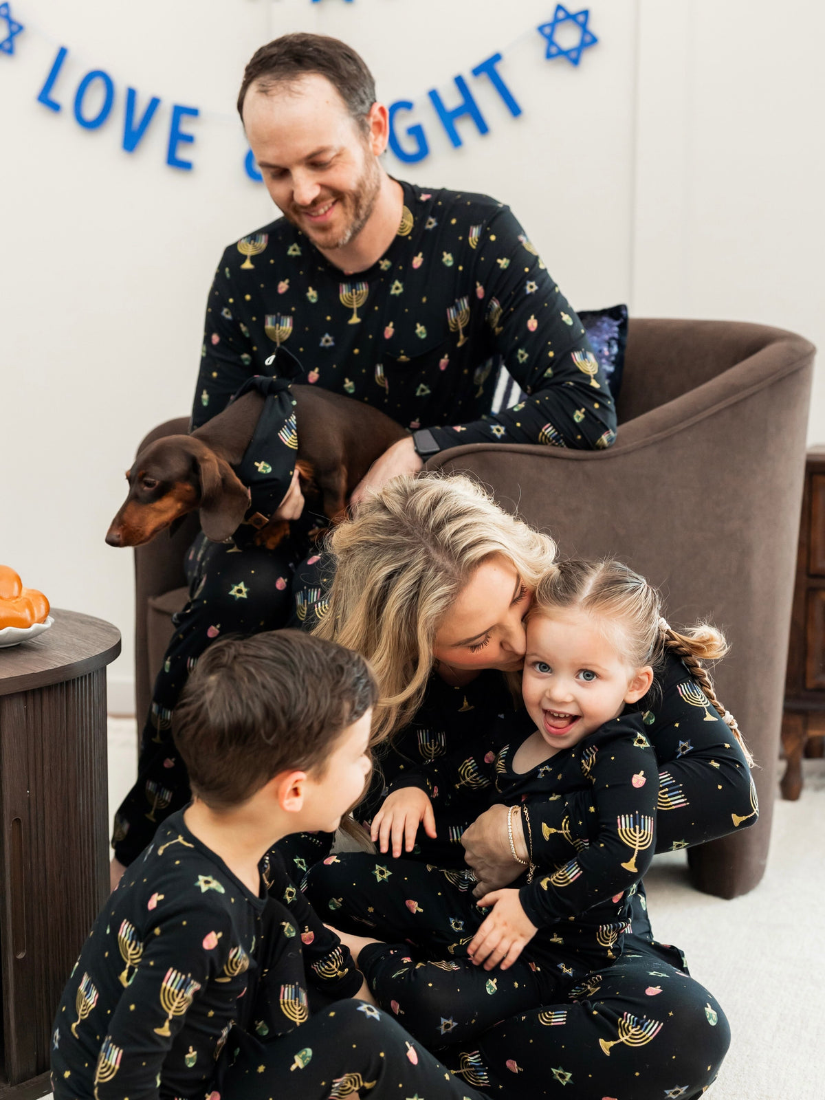 A family of four wearing matching black pajamas with a colorful menorah and Star of David pattern, gathered in a cozy living room setting with a brown armchair and a dachshund.