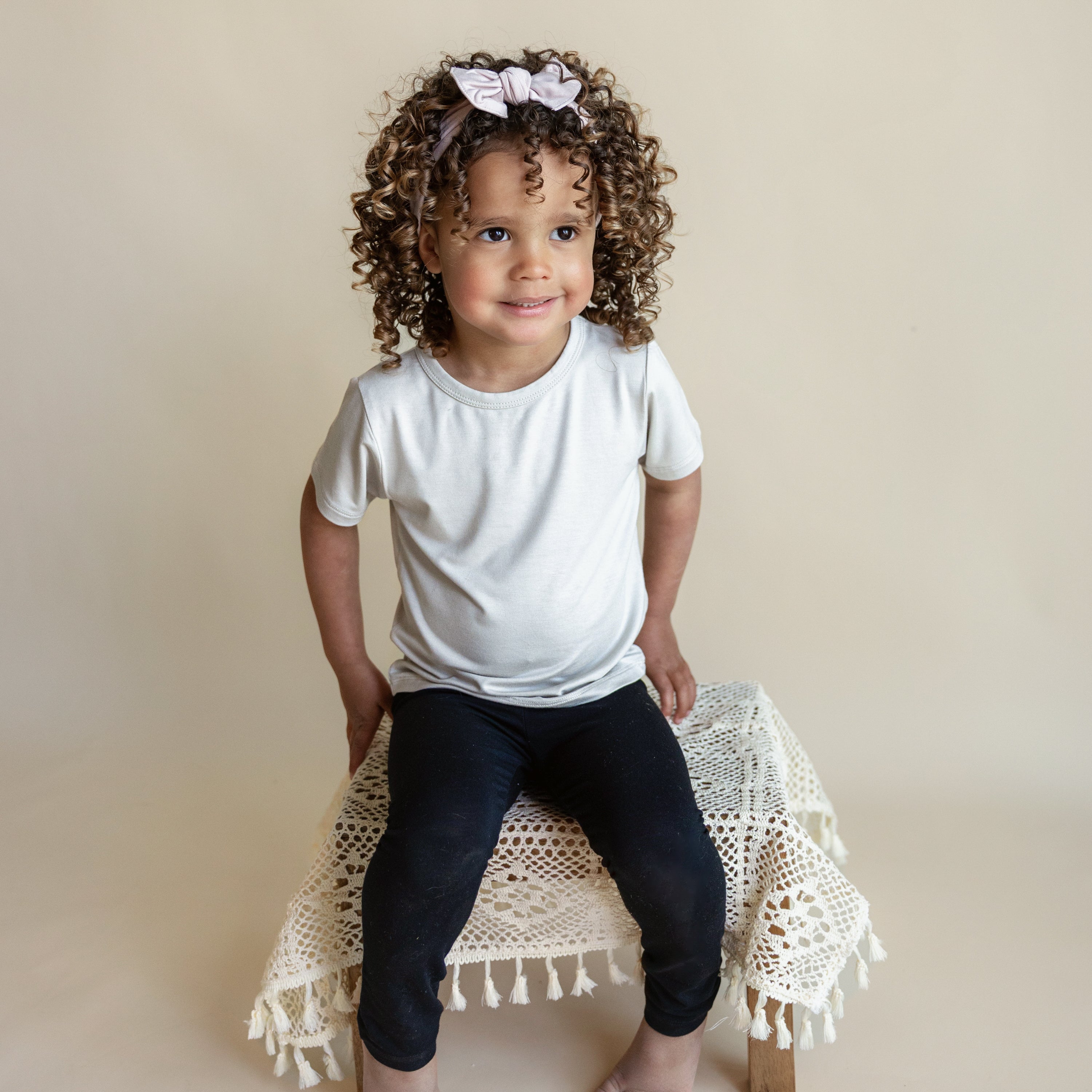 Smiling toddler sitting on a wooden stool draped with a cream crochet cover while wearing Toddler Leggings in Midnight and a Tee in Oat