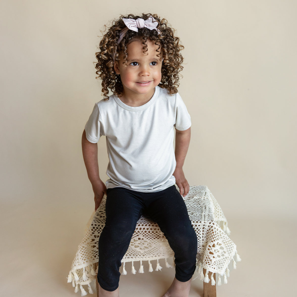 Smiling toddler sitting on a wooden stool draped with a cream crochet cover while wearing Toddler Leggings in Midnight and a Tee in Oat
