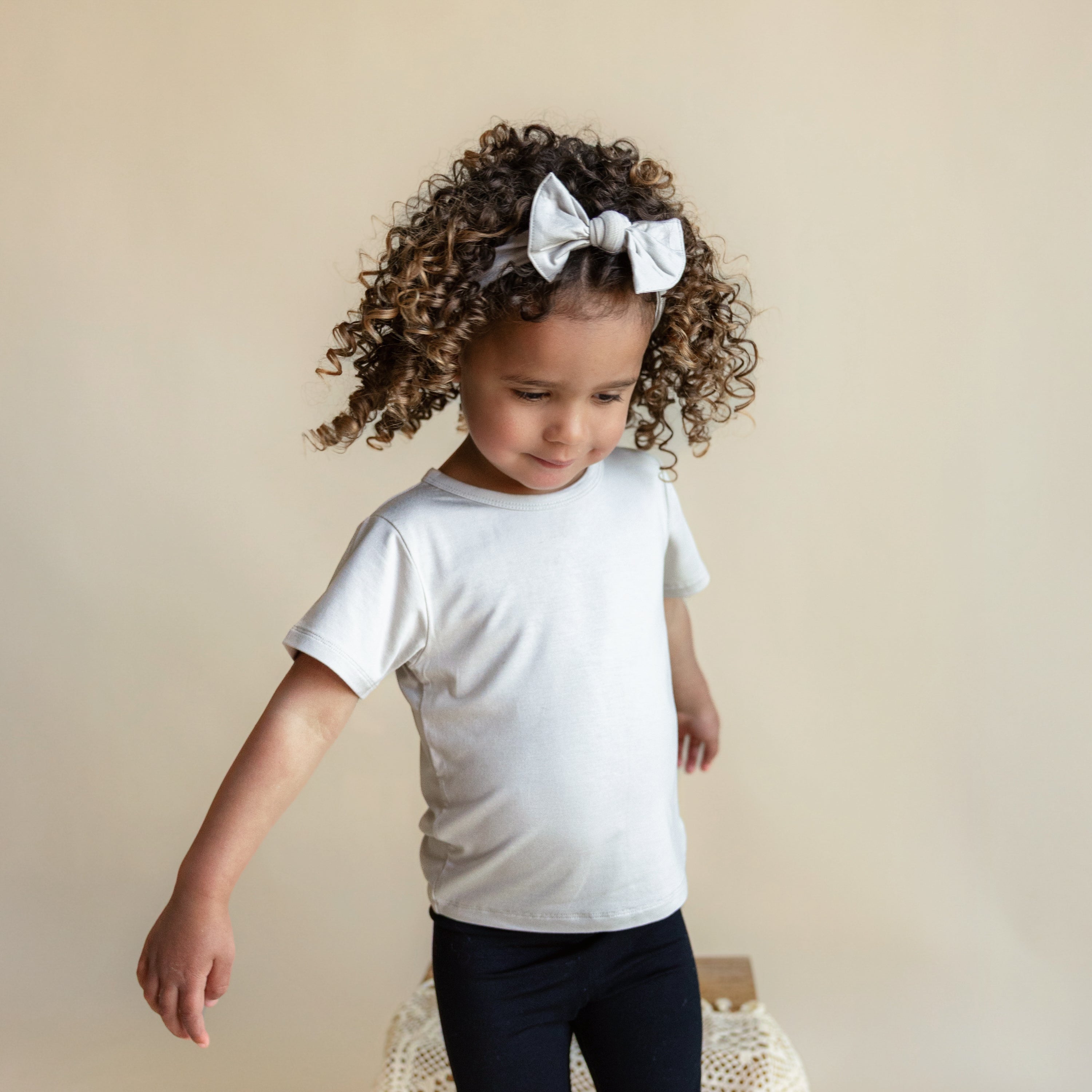 Little girl wearing Knotted Bow Headband in Oat while standing in front of a beige wall