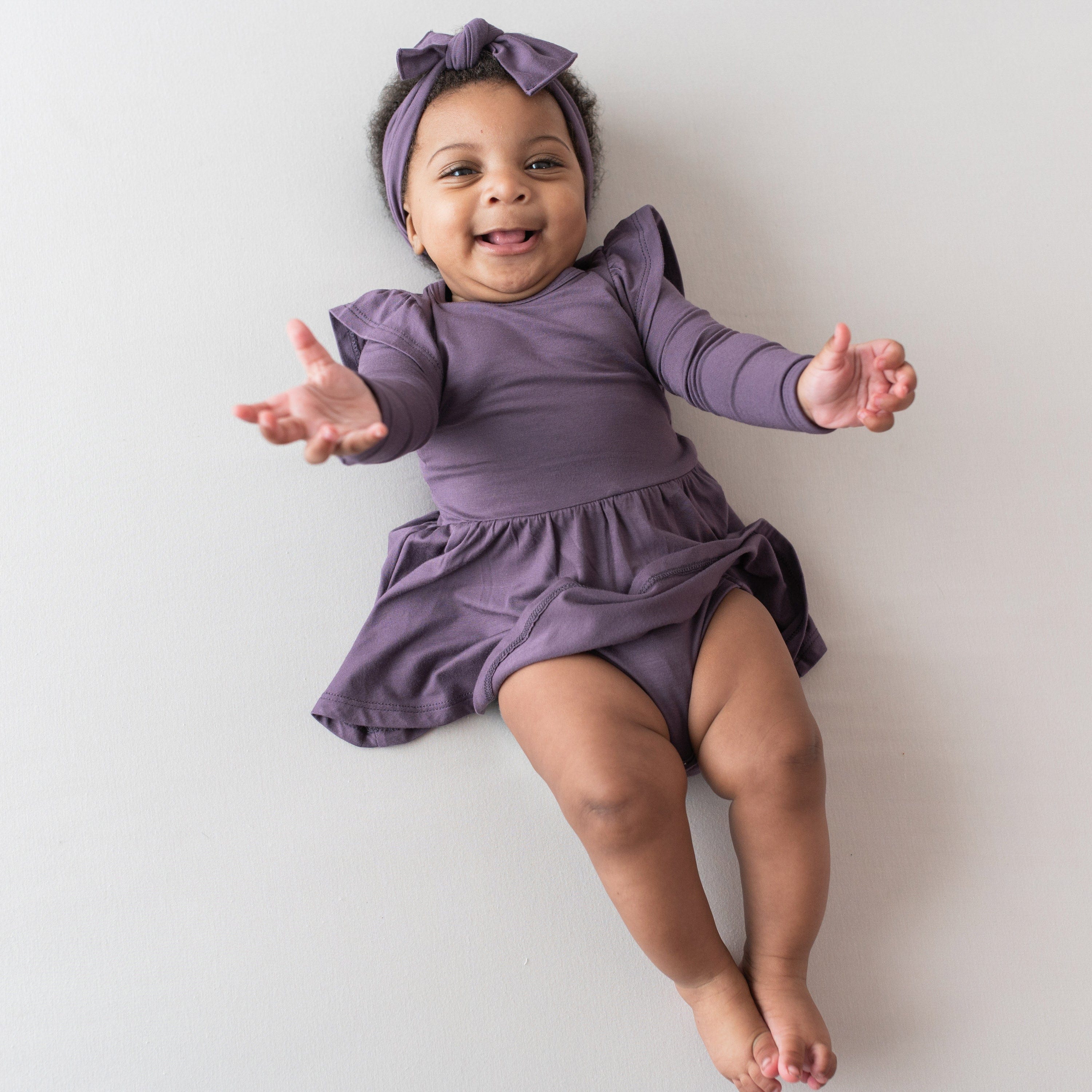Infant wearing Kyte Baby Twirl Bodysuit Dress in Currant with purple bow headband