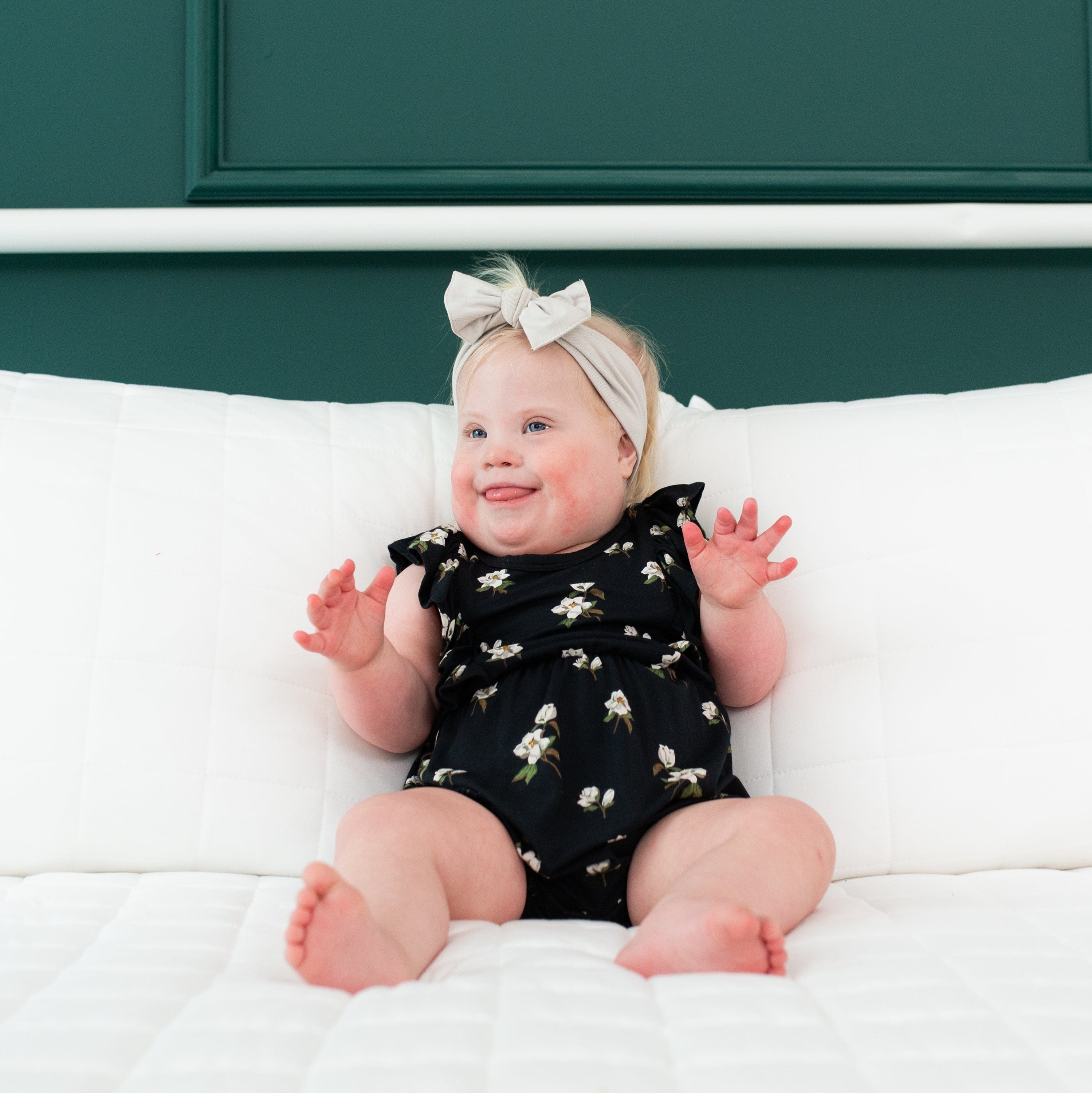 Baby sitting on a white bed while wearing a Bow in Oat and a Midnight Magnolia Bubble Romper