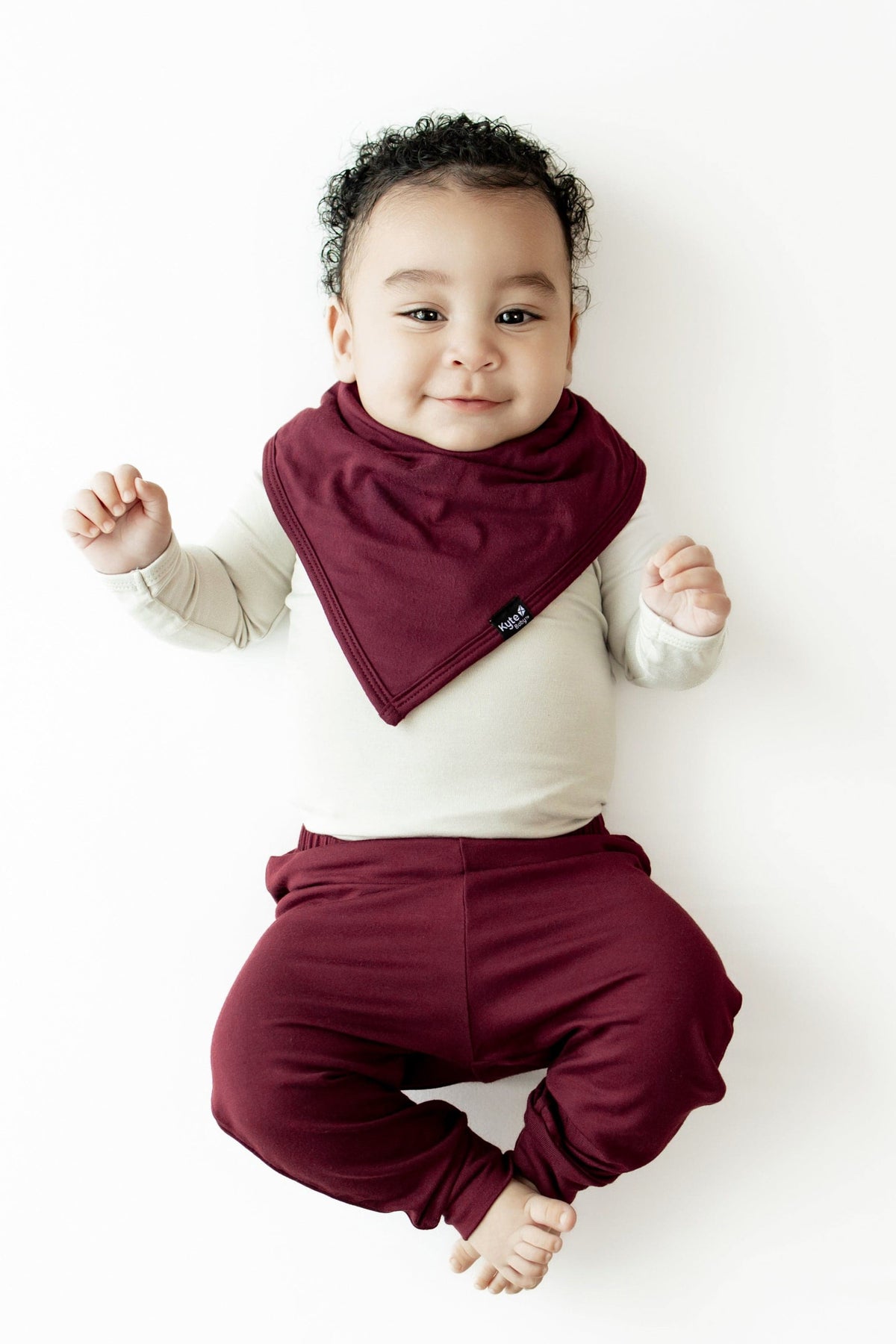A baby lying on their back wearing a burgundy triangle bib over a cream long-sleeve top and matching burgundy pants, photographed against a clean white background.