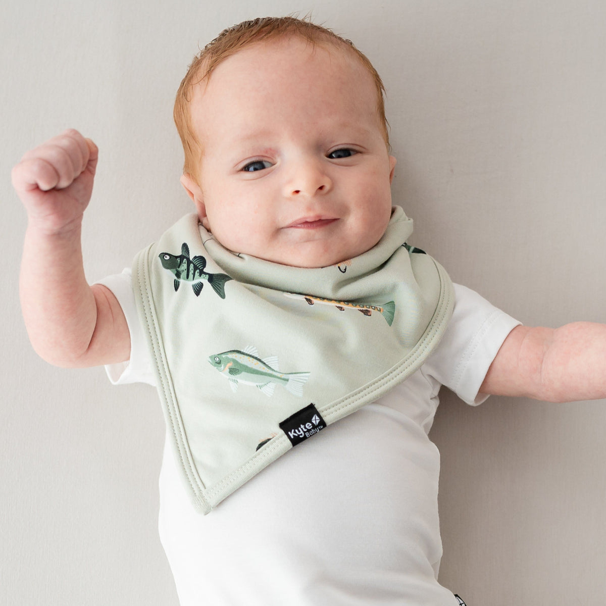 Infant laying down on a light neutral surface wearing the Bib in Fishing and a white bodysuit