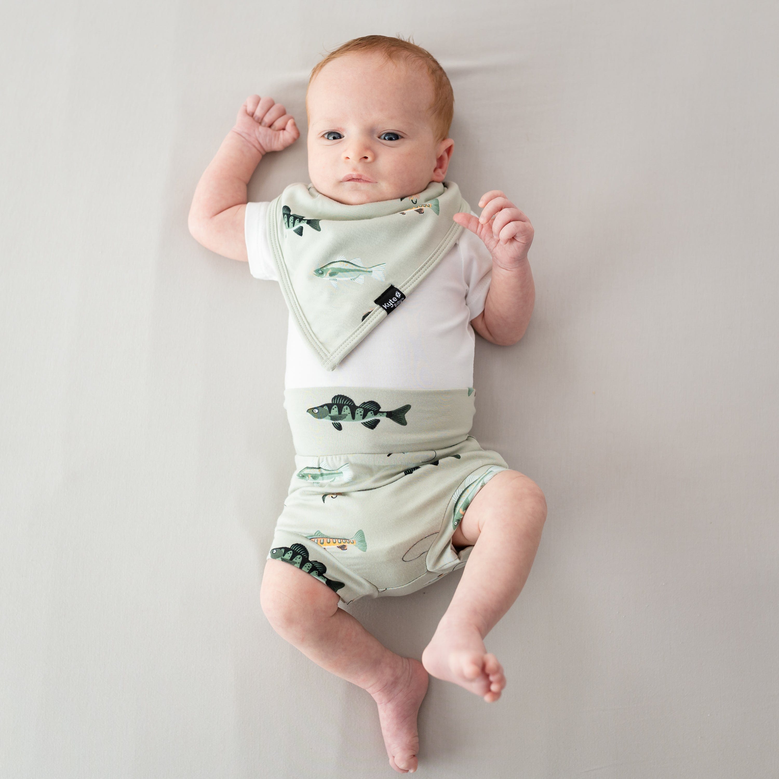 Infant laying down on a light neutral surface waring the Bib in Fishing with matching bummies and a white bodysuit