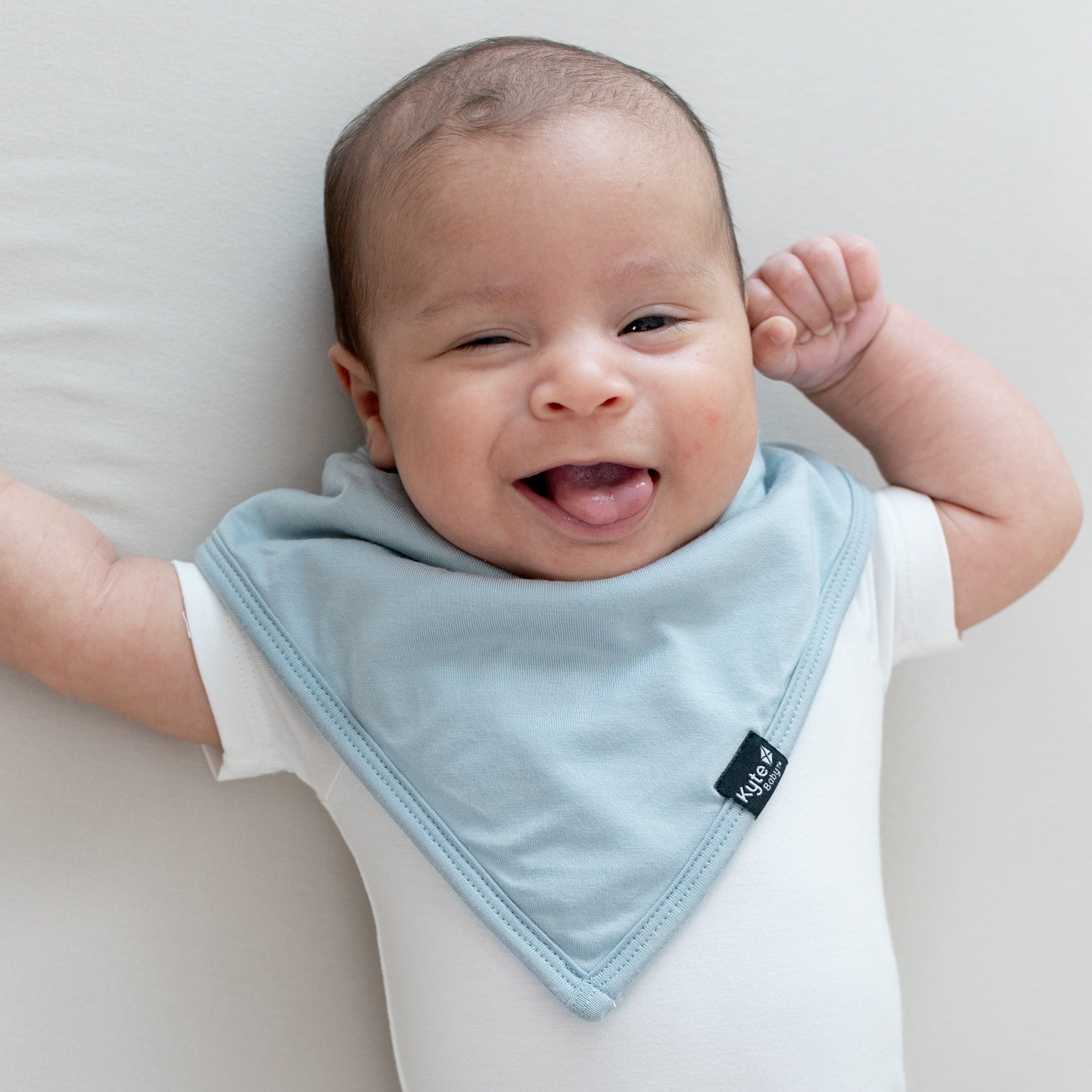 Infant laying down sticking out their tongue wearing the Bib in Fog and a white short sleeve bodysuit
