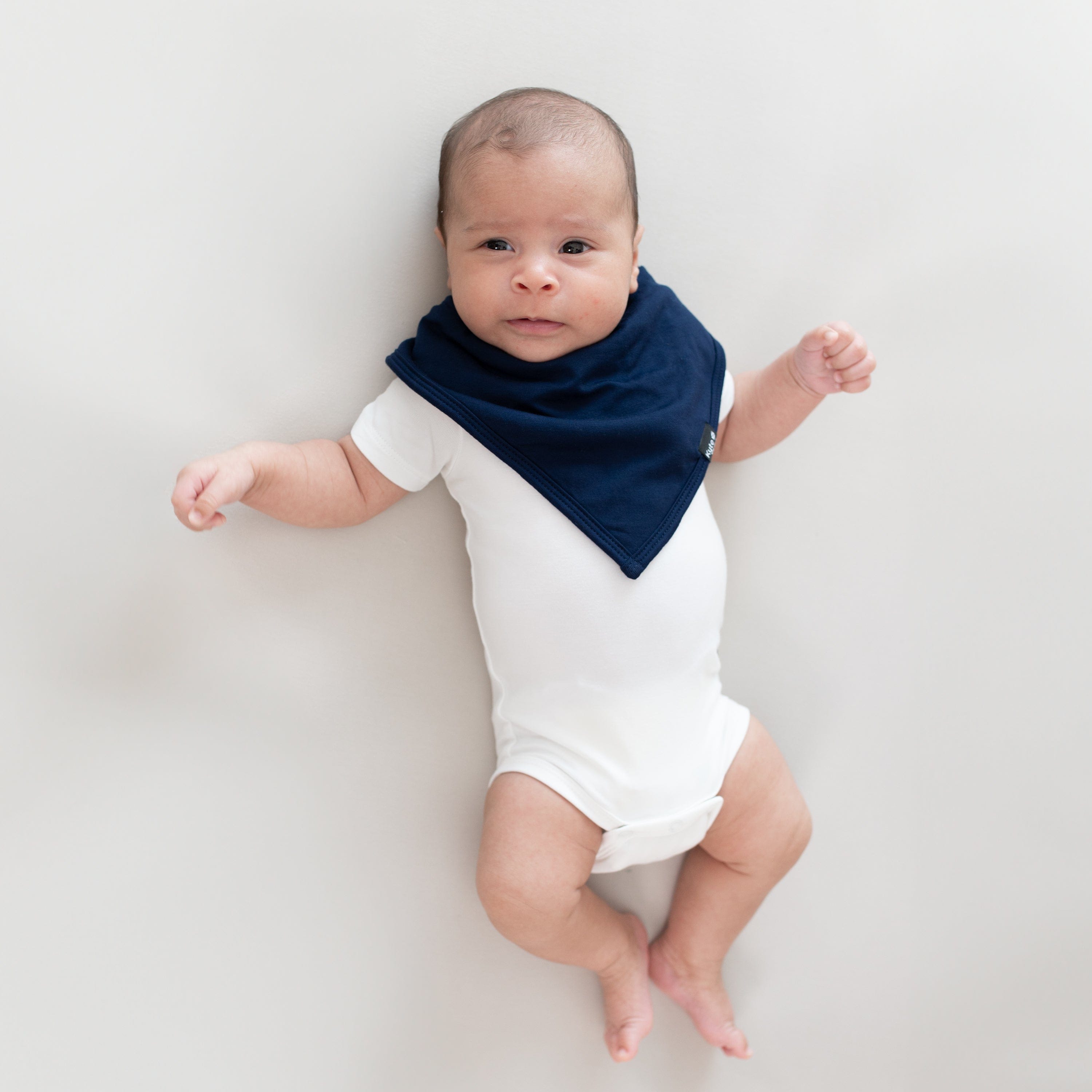 Infant laying on a Crib Sheet in Oat while wearing a Bib in Navy