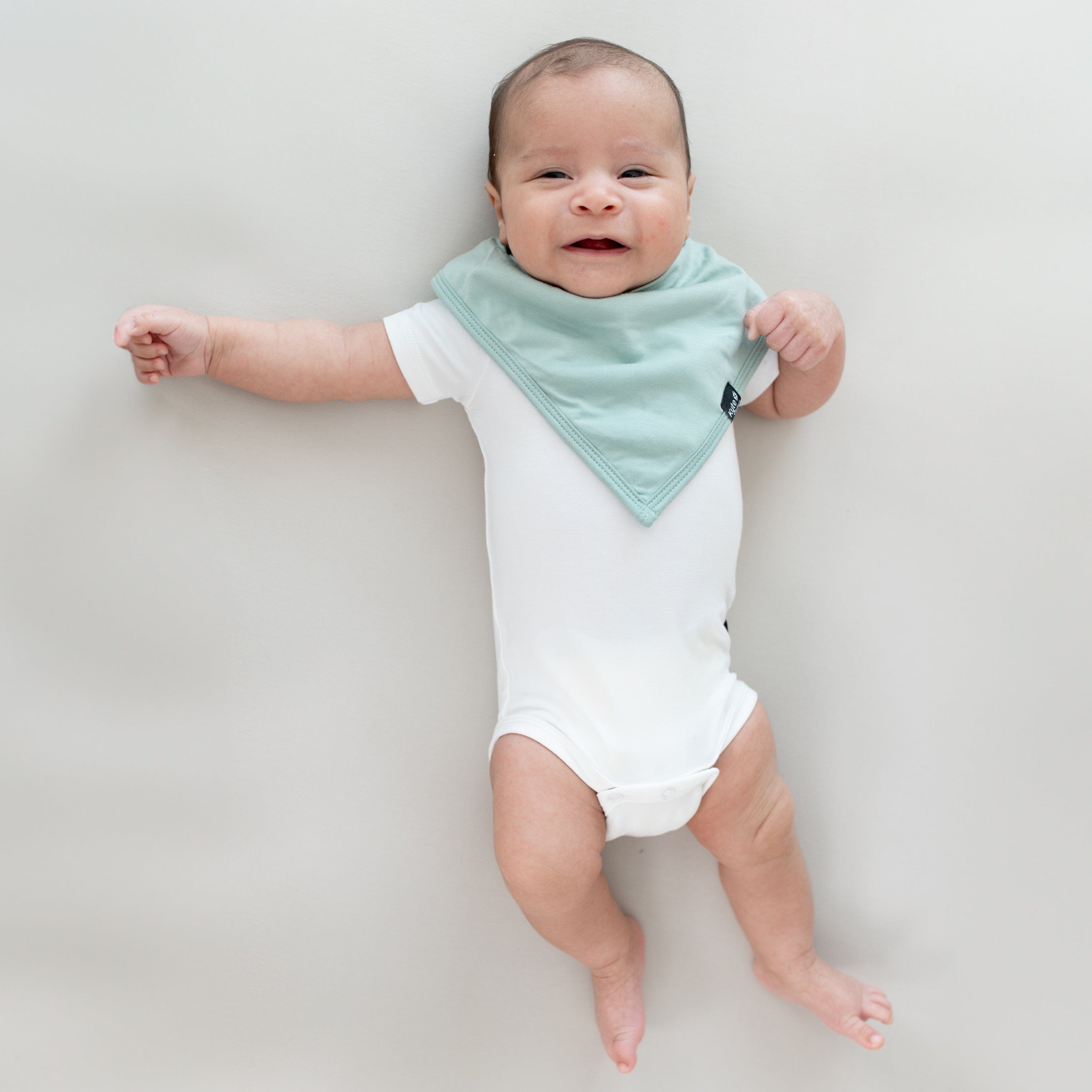 Baby laying on a Crib Sheet in Oat while wearing a Bib in Sage