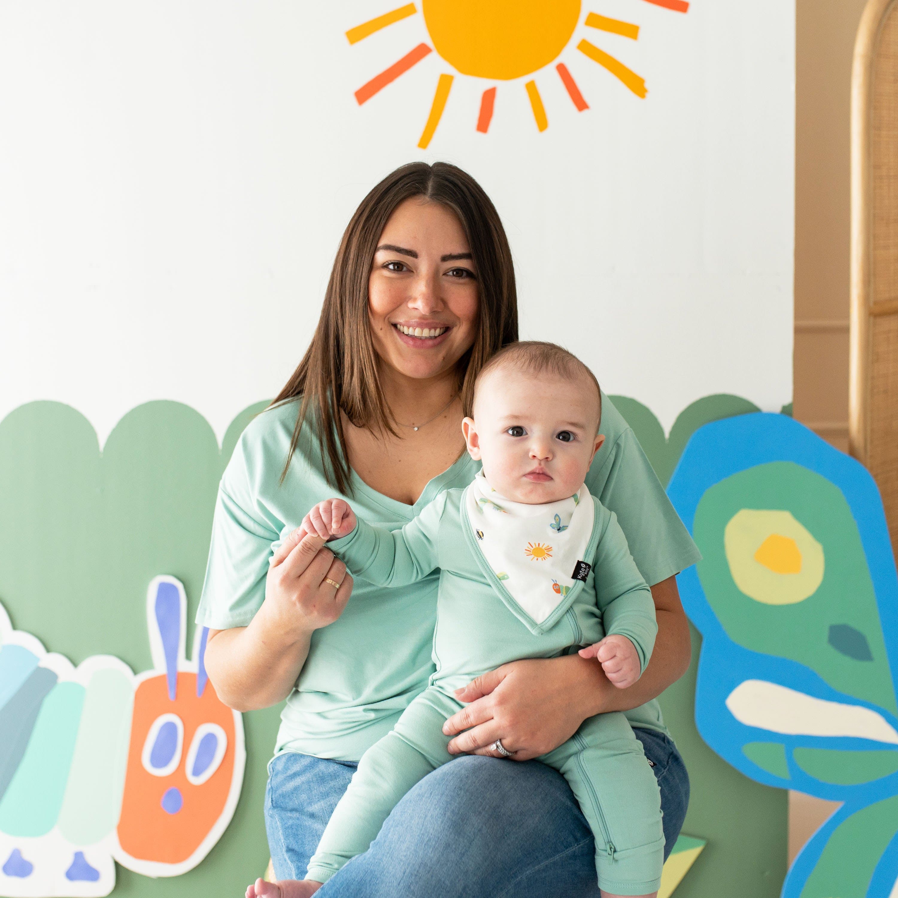 Mother wearing a green shirt holding her son who is matching and wearing a Bib in The Very Hungry Caterpillar™ and Friends