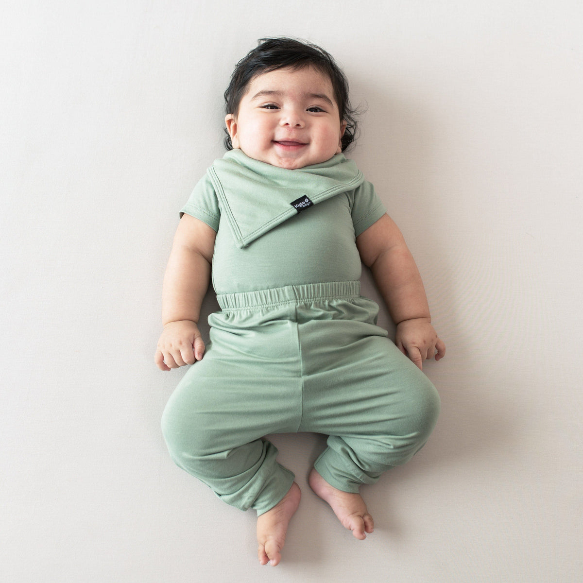 Infant model laying on a beige surface wearing the Bib in Thyme and matching short sleeve bodysuit and pants