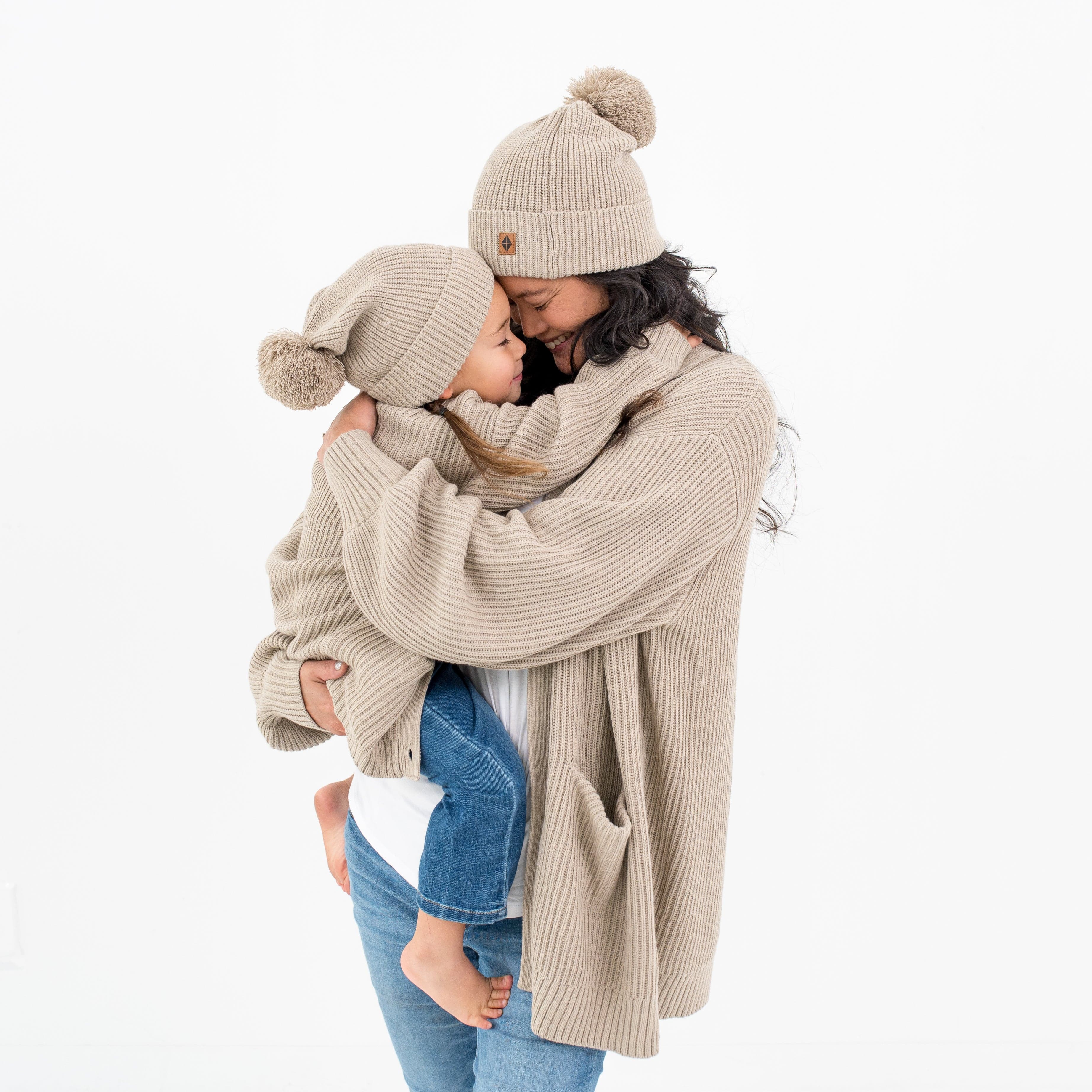 Mom and child hugging in Kyte Baby Chunky Knit Adult Pom Beanies in Almond